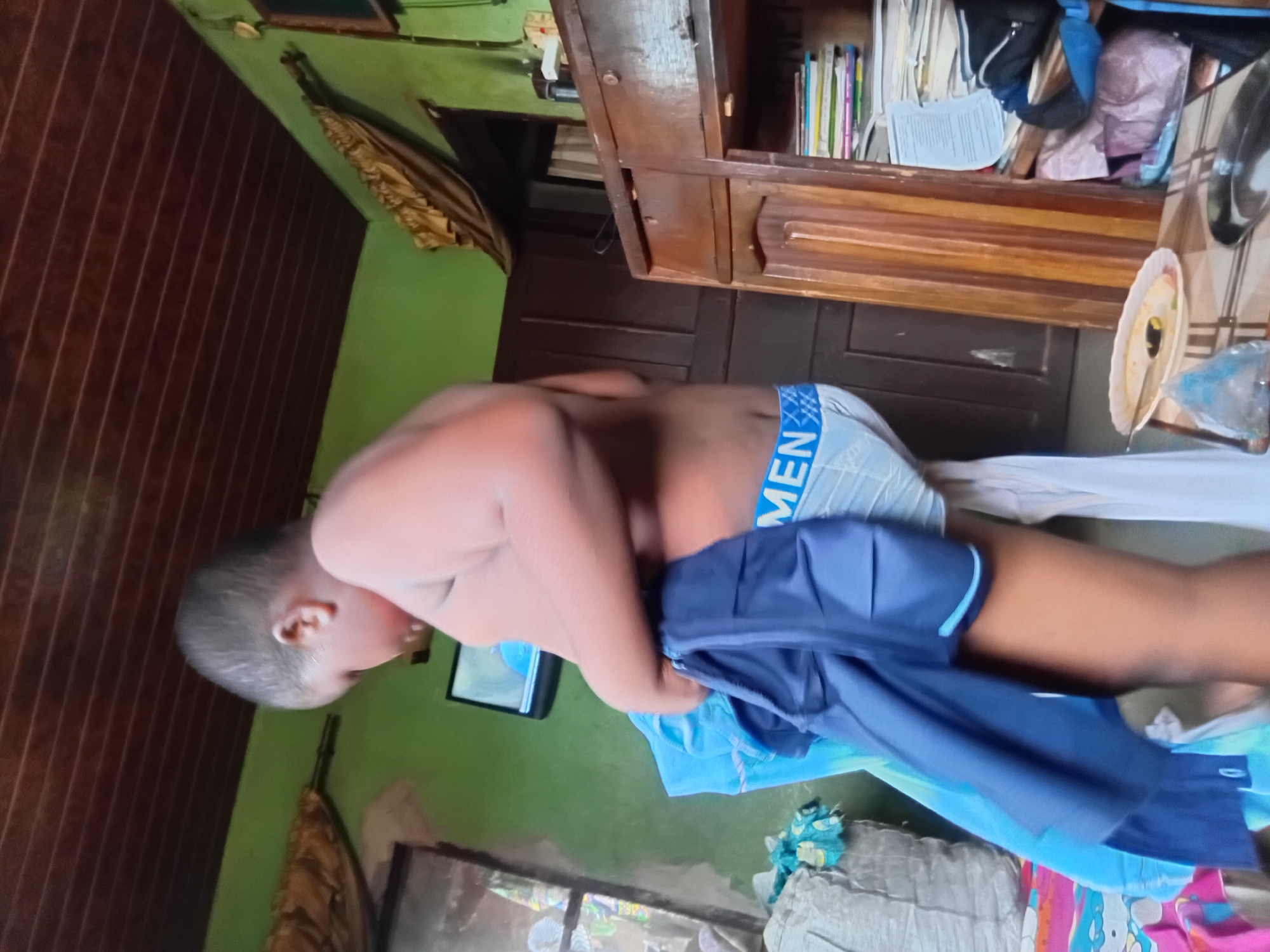 A young boy, possibly a toddler, is captured in a vertical, dimly lit indoor shot. He is wearing light blue underwear with the word "MEN" visible in blue text. His body is turned away from the camera, with his head tilted down. He is holding a dark blue garment, possibly shorts, which obscures part of his body.
The setting appears to be a room with green painted walls. To the left, a wooden paneled wall is visible. In the background, a wooden cabinet with shelves filled with books and other items stands against a dark wooden wall. A small television screen is visible on a shelf in the background, showing a blurry image. The floor is not clearly visible.
The lighting suggests it could be daytime, but the indoor setting and shadows make it difficult to determine the exact time of day. There are no clear indicators of weather or specific emotional states, although the boy's posture might suggest he is playing or changing. The location context of Nkpor, Nigeria is provided, but no explicit visual cues confirm this. Overall, the image captures a candid moment of a child in a domestic environment.