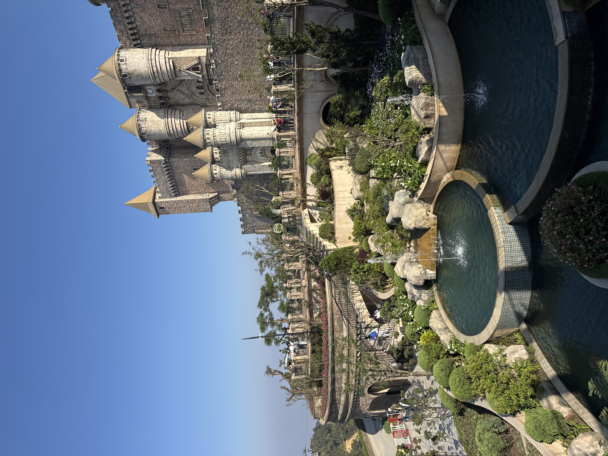 The image captures a detailed panoramic view of Sun World Ba Na Hills, specifically the French Village, situated in the Truong Son Mountains west of Da Nang, Vietnam. The scene features a large, meticulously constructed European-style stone castle occupying the upper right and central background, characterized by multiple turrets, battlements, pointed golden spires, and a visible clock face on a prominent tower.
Below the castle, an elaborate terraced landscape unfolds, incorporating manicured gardens with diverse green foliage, shrubs, trees, and flowering plants in shades of white and pink/purple. Interspersed within these gardens are multi-tiered water features, including several interconnected pools with blue mosaic tiling, from which multiple fountains actively spray water upwards. Natural rock formations are integrated into the garden and around the water bodies.
Architectural elements include stone arches supporting bridges and walkways, and decorative black metal railings line the elevated pathways and balconies. Numerous individuals are visible across the complex: groups are present on a main pedestrian bridge in the midground, on a higher curved balcony to the left, and on a lower paved area near red and white barriers on the far left. A few individuals are also seen descending a set of stairs. A cable car line, with a gondola, is faintly discernible in the upper left background, indicating the mountainous terrain. The sky is clear and blue, and the overall lighting suggests bright daylight conditions, with a slight atmospheric haze visible in the distant background on the left.