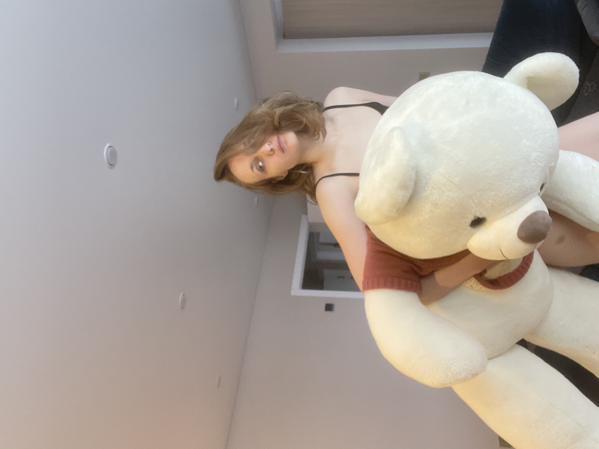 An indoor, low-angle photograph captures a young woman holding a large plush toy. The woman, with shoulder-length light brown hair, is seen from the chest up, wearing a black bra or camisole. She is looking directly towards the camera with a neutral expression, showing a faint hint of a smile. Her left arm is visible, holding the significant weight of the plush toy.
The plush toy is a large, cream-colored bear, strongly resembling Winnie the Pooh, and is wearing a rust-red knitted sweater with a lighter yellow or cream trim around the neck. The bear's face features small, dark eyes and a brown nose.
The setting is a room with light-colored, possibly white or off-white, walls and ceiling. The perspective is from below, looking up towards the subjects and the ceiling. Two circular recessed light fixtures are visible on the ceiling. A rectangular opening, which could be an attic access, a skylight, or a vent, is visible in the upper background. A dark object or piece of furniture is partially visible in the far upper right corner of the frame. The lighting suggests an indoor daytime setting. The image provides a context of being taken in Подшивалово, Russia. No visible text is present in the image.