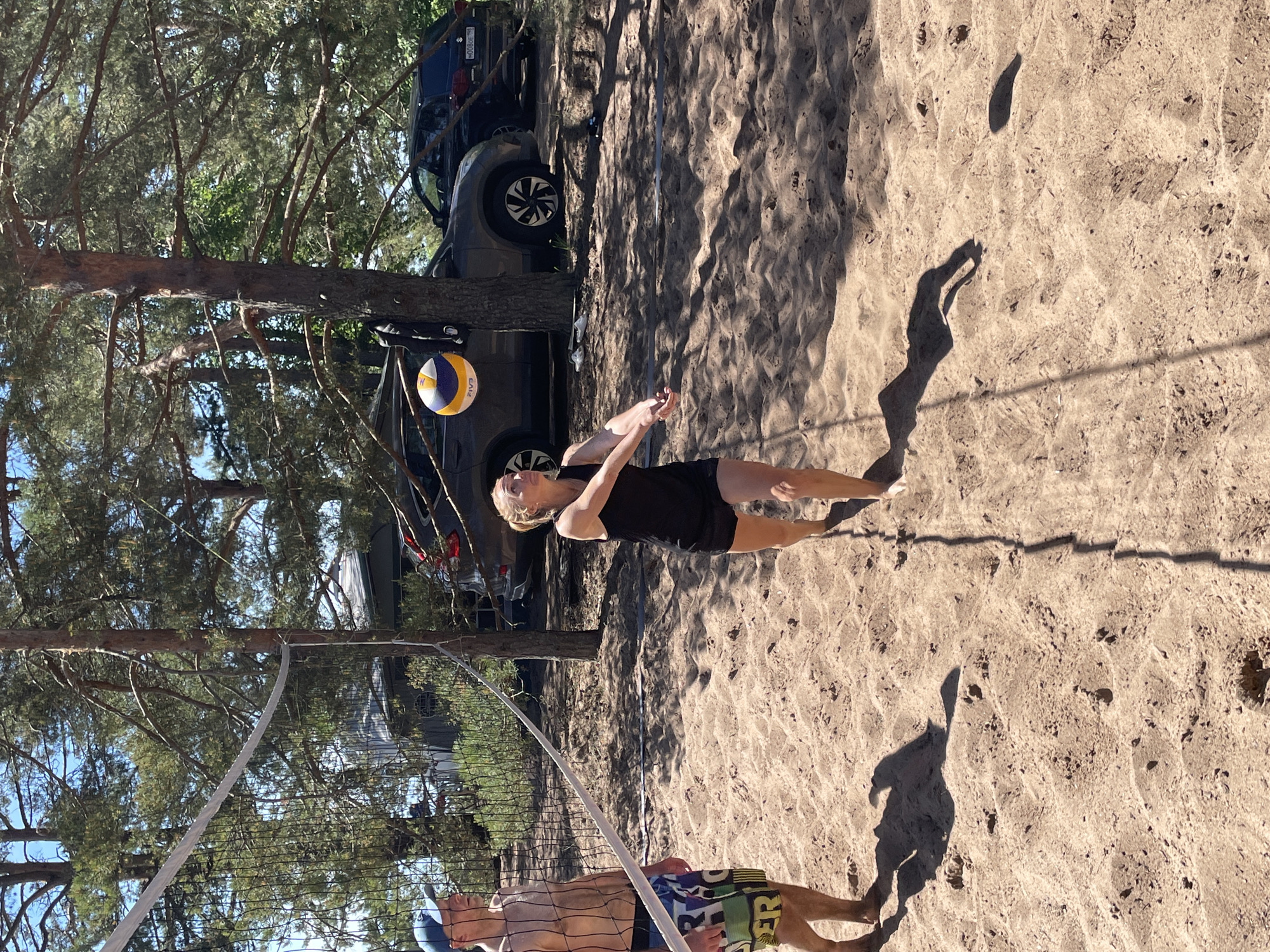 The image depicts an outdoor beach volleyball scene in Zelenaya Roshcha, Russia, under clear blue skies and bright daylight. The ground is covered in light brown sand, marked with footprints and shadows.
In the foreground, a young adult female with light-colored hair, wearing a black tank top and black shorts, stands barefoot. She is positioned with her arms extended forward, looking upward at a volleyball, indicating she is preparing to execute a bump pass. A multi-colored (blue, yellow, white) "FIVB" branded volleyball is suspended in the air slightly above and to her right.
To the left, separated by a white-banded black volleyball net, stands an adult male. He is shirtless, wearing a blue baseball cap and colorful patterned swim trunks (blue, green, yellow, white) with "ER" visible on one leg. He is also barefoot and looking towards the right, observing the female player and the ball.
The volleyball net is strung between two points, one of which is a prominent, dark-barked pine tree trunk on the right side of the frame. Numerous other tall pine trees with green foliage form a dense canopy in the background, casting dappled shadows across the sand.
Several vehicles are parked behind the trees in the mid-ground. A dark grey SUV is visible behind the net, with a yellow and black item hanging from the tree trunk beside it. Further to the right, a darker-colored SUV (likely a Honda CR-V) is partially visible, showing its rear light and a Russian license plate "M008OE 198". Parts of buildings or structures are vaguely discernible through the trees in the far background.
