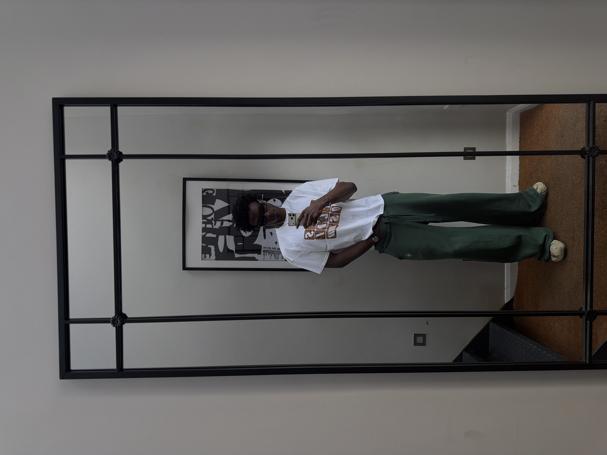 A young Black man is taking a mirror selfie in what appears to be a residential hallway in London, United Kingdom. He is standing in front of a large, multi-paneled mirror with a black frame. The man is wearing a white t-shirt with a graphic print, dark sunglasses, dark green wide-leg sweatpants, and light-colored sneakers. He is holding a smartphone in his right hand, and his left hand is in his pocket.
The mirror reflects the man, a framed piece of black and white abstract art hanging on the wall behind him, and a staircase with dark metal treads and a black railing to the left of the frame. The floor in front of the mirror is covered with a brown carpet. The wall on the right of the frame is a light beige color and shows some subtle texture. A black electrical outlet is visible on the wall behind the man, and a small square light switch is visible on the wall to the left of the frame. The lighting in the room is neutral, suggesting it is daytime and natural light is supplemented by artificial lighting. The overall impression is one of a casual, modern interior space.