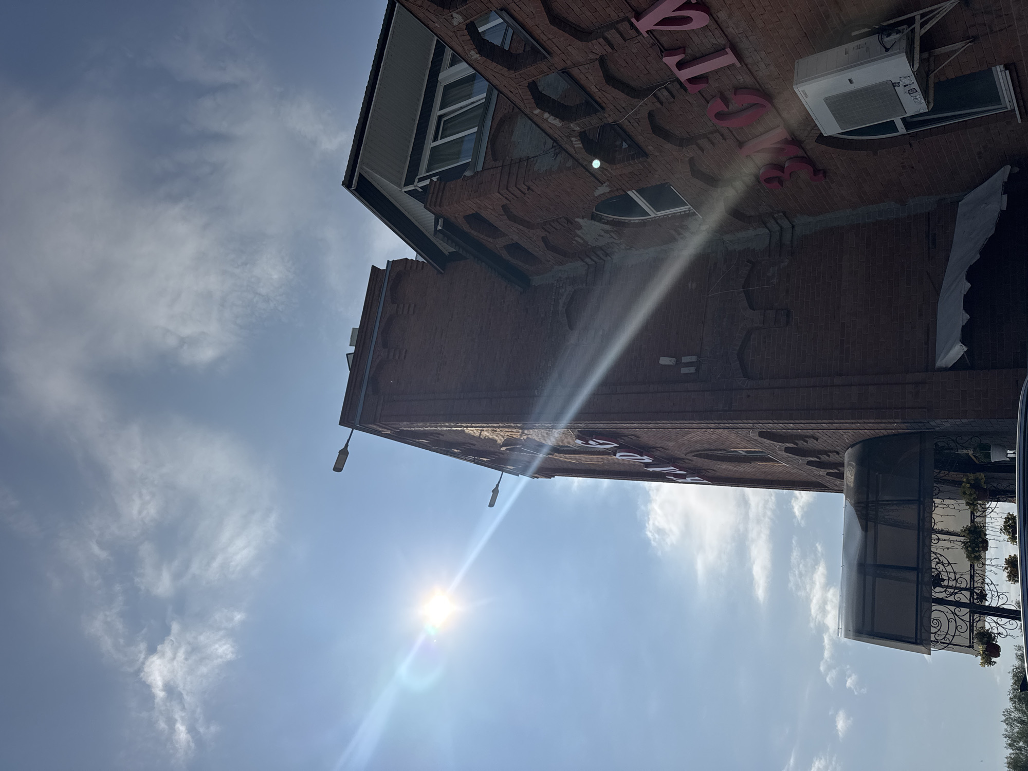 The image shows a brick building with a sign that appears to read "ЗАСТАВА" in pink lettering. There are arched windows and what looks like an air conditioning unit mounted on the side of the building. A glass awning with decorative metalwork extends from the building over what might be an entrance. The sun is bright and high in the sky, creating lens flare and casting a bright ray across the scene. The sky is blue with scattered white clouds. In the distance, there are trees. The building is located in Verkh-Sosnova, Russia.