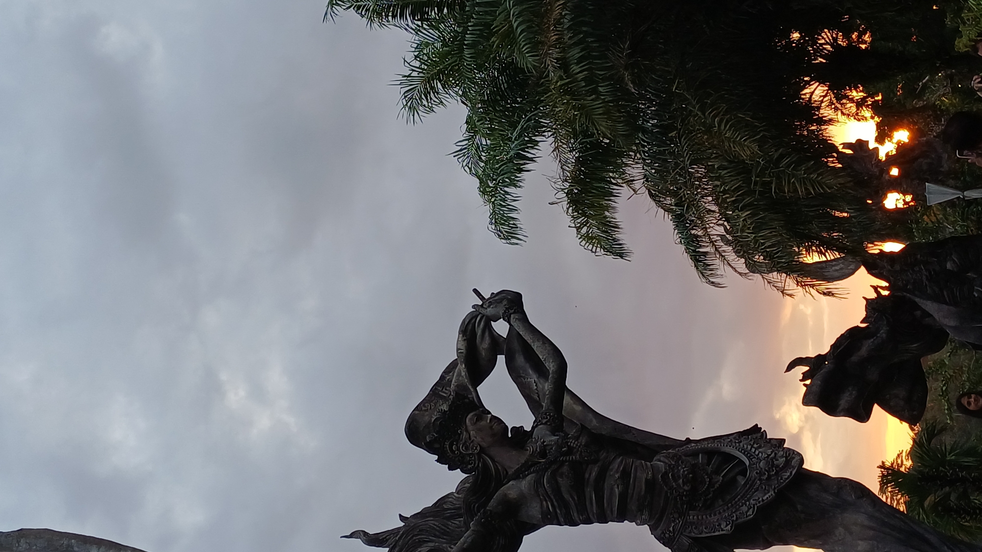 The image shows a low-angle view of a statue in Ubud District, Indonesia, set against a sky that blends from grey to orange hues. The statue appears to be of a human figure with wings, raising an object in its hands, possibly a musical instrument or a ceremonial object. The statue is intricately detailed, showing ornate carvings and drapery. To the upper right of the frame, there are palm trees silhouetted against the bright orange sky, hinting at a sunset. Another statue, possibly of a bull or similar animal, is partially visible on the right side, contributing to the cultural or religious significance of the location. The overall environment suggests an outdoor setting, likely a temple or a significant landmark in Ubud.