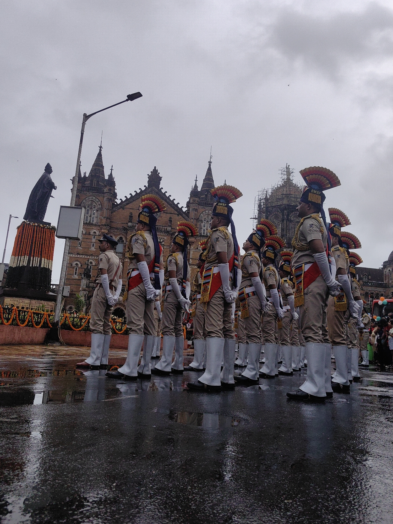 This is a full-shot, eye-level, outdoor photograph taken in Mumbai, India, likely at the Chhatrapati Shivaji Maharaj Terminus, a UNESCO World Heritage Site, given the architectural style of the building visible in the background. The image captures a group of approximately 20 individuals dressed in formal tan uniforms with white boots, white gloves, and distinctive red and yellow feathered headwear. They are standing in formation on a wet, reflective paved surface, suggesting recent rain. The uniformed individuals are facing to the left of the frame, appearing to be participating in a parade or ceremony. Behind them, the ornate, gothic-Victorian architecture of a large building is prominent, featuring spires and detailed stonework. To the far left of the frame, a dark statue of a man in a robe stands on a pedestal adorned with garlands of orange and white flowers. The sky is overcast and a dull grey, with a few small birds visible. In the background, scaffolding can be seen on top of one of the building's towers, and a crowd of onlookers dressed in casual clothing is visible on the far right. The scene evokes a sense of formal occasion against a backdrop of historical architecture in a public space.