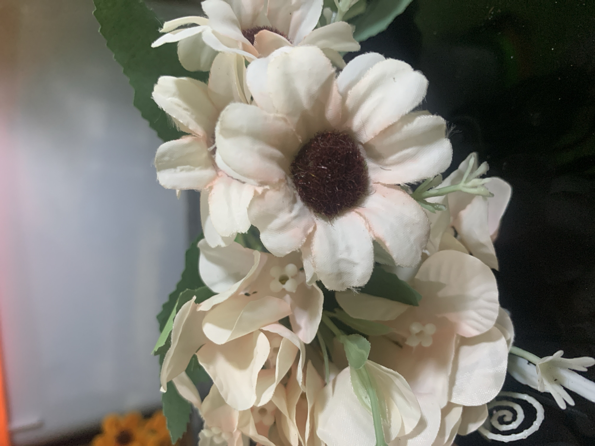 The image is a close-up of a bouquet of artificial flowers. The flowers are predominantly pale pink and cream with some green leaves interspersed. The focus is on the textured petals and the dark brown centers of some of the flowers, which resemble daisies. There is a hint of other flowers in the background, one with a bright orange hue and a sunflower-like appearance. The bouquet is resting on a dark, reflective surface, possibly a table. The background is out of focus, showing a blurred gradient of light grey to white, with a sliver of an orange light source at the top. The overall impression is of a still life arrangement.
There is no indication of Kuala Pembuang, Indonesia, or any specific location within a city. The image appears to be a studio or indoor shot of decorative items.