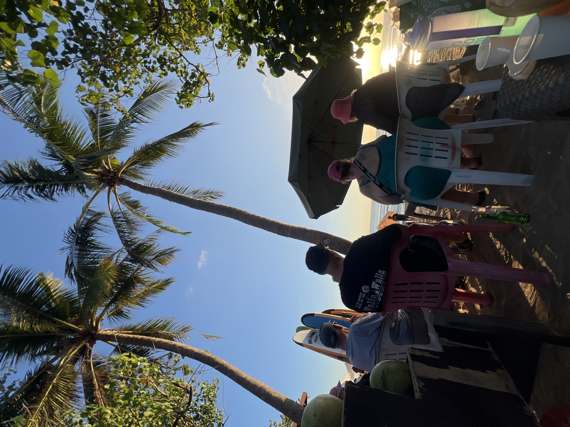 In Kuta, Indonesia, four individuals are seated on the beach, facing the ocean at sunset. Two palm trees frame the scene, casting shadows on the sandy ground. In the foreground, a wooden structure partially obscures a person from the chest down, sitting on a pink chair. To their left, another person sits on a white chair. In the middle ground, two individuals, a woman in a teal dress and a person in a dark jacket, are seated under a large green umbrella. Behind them, surfboards stand upright against a beachside establishment. The ocean is visible in the distance, with the sun setting on the horizon, casting a golden glow on the water. Several coconuts and drinks are scattered around the seating area, suggesting a relaxed atmosphere at a beachside bar or cafe.