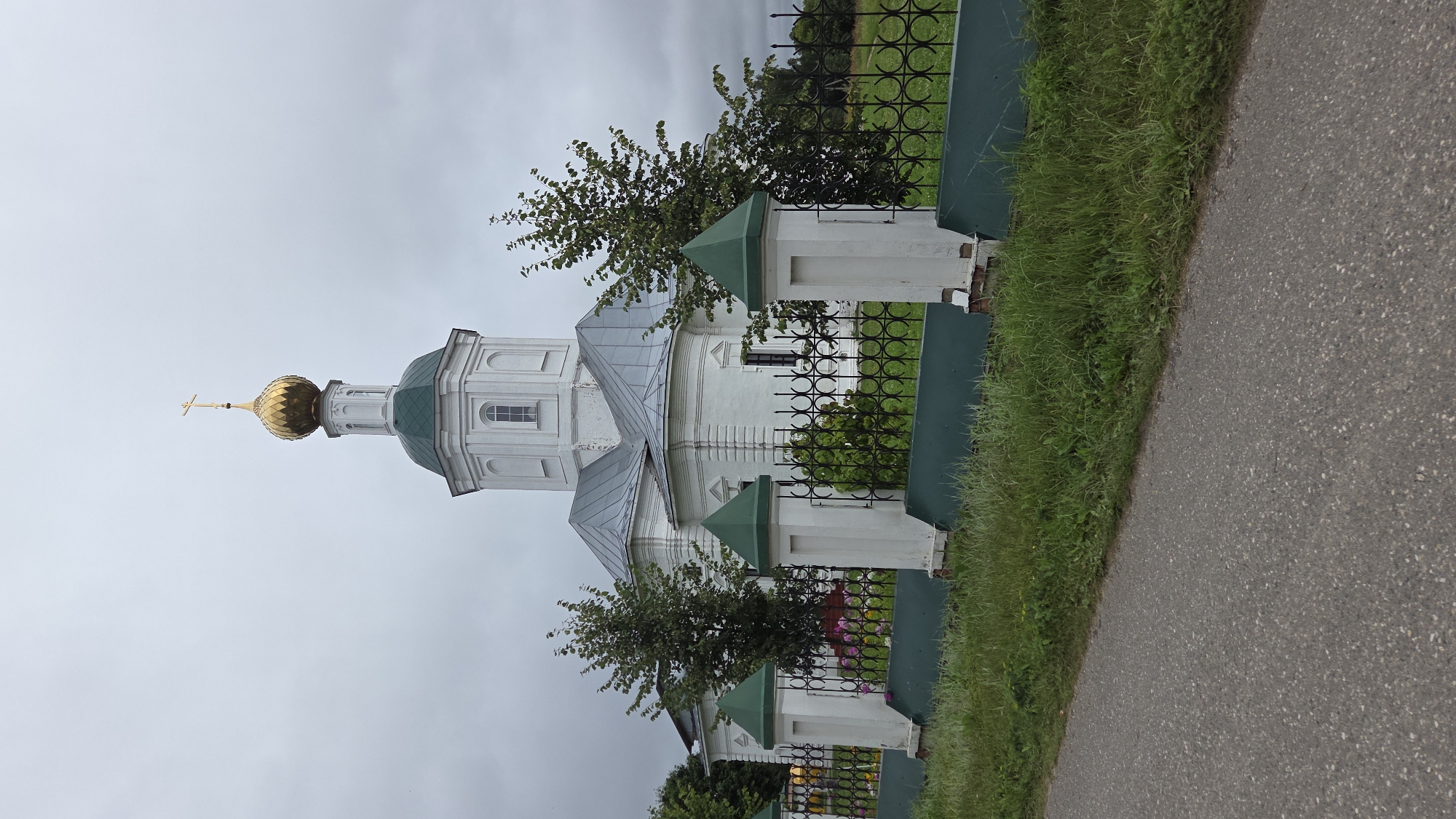 The image shows a white orthodox church with a green roof and a golden dome topped with a cross. The church is surrounded by a black metal fence with white pillars topped with green pyramid-shaped caps. A paved road is visible in the foreground, with grass and some flowers behind the fence. Trees are scattered around the church. The sky is overcast. The church is located in the city of Novo-Talitsy, Russia.