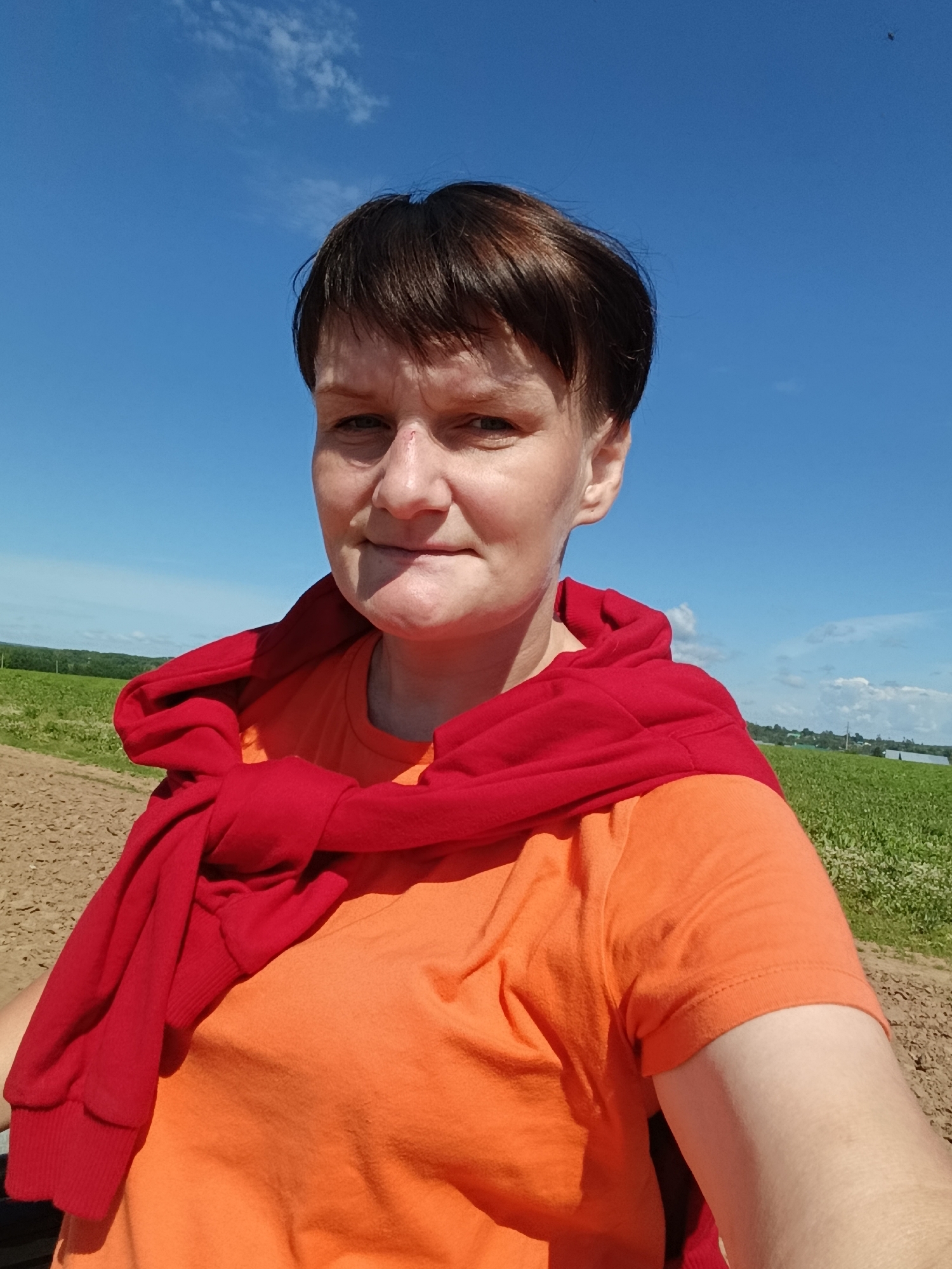 The image is a close-up selfie taken outdoors under a clear blue sky with scattered clouds. The subject is a woman with short, dark brown hair, and fair skin. She is wearing an orange t-shirt with a red sweater tied around her shoulders. The background shows a field with some crops, possibly agricultural land, and a dirt path in the foreground. The location is identified as Fokino, Russia. The overall scene depicts a casual, outdoor setting.