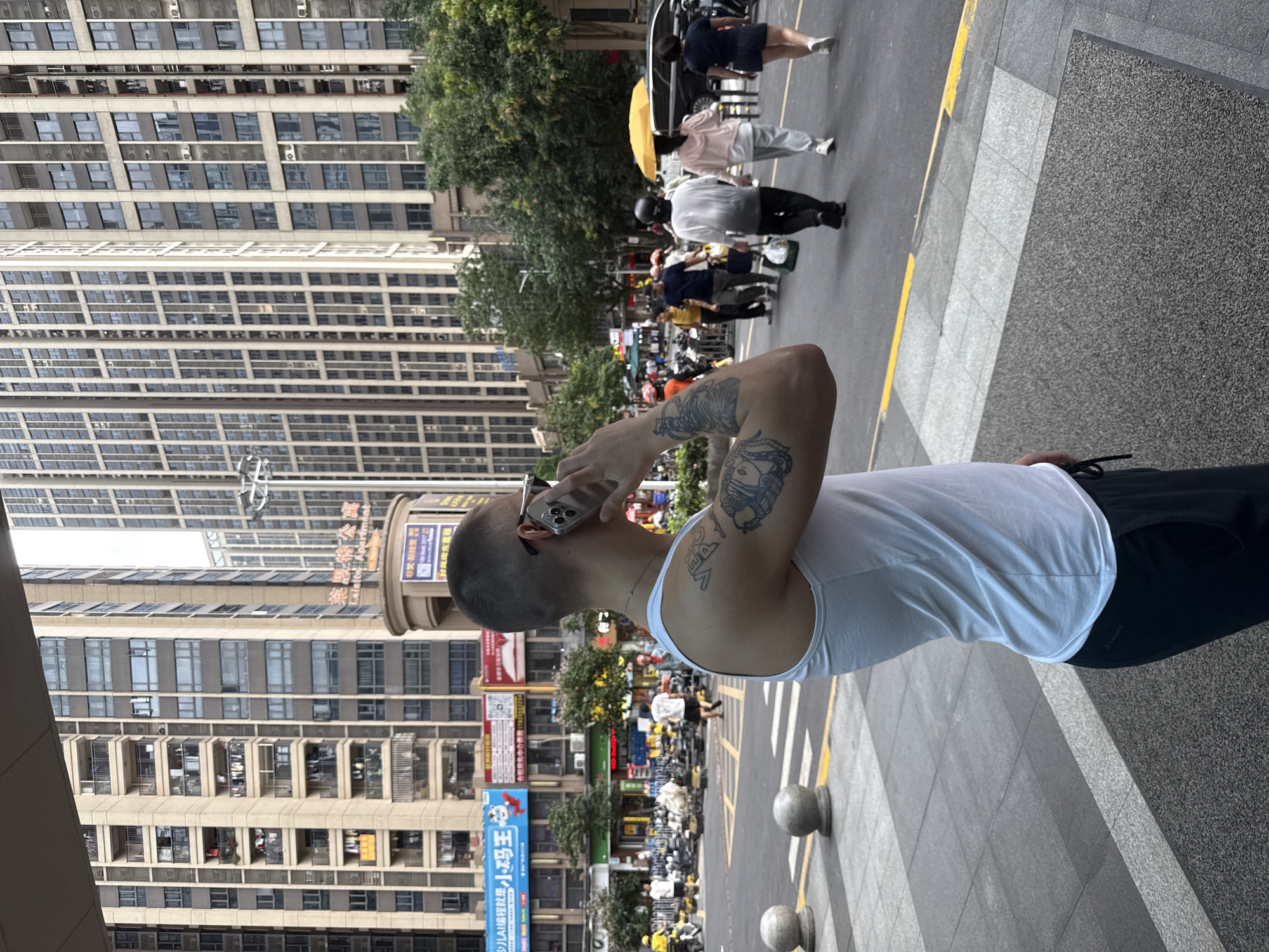 The image captures a street scene in Haizhu District, China, focusing on a man in the foreground speaking on his phone. He is wearing a white tank top and black shorts, with visible tattoos on his arm. The man is positioned on a sidewalk, with a street and a busy intersection behind him. Several pedestrians are visible in the background, some walking, others on scooters or motorbikes, suggesting a typical urban environment. Tall apartment buildings with numerous balconies and windows dominate the cityscape, indicative of a densely populated area. The presence of shops and signage suggests commercial activity. The time of day appears to be daytime, with the lighting suggesting it could be late afternoon or early evening.