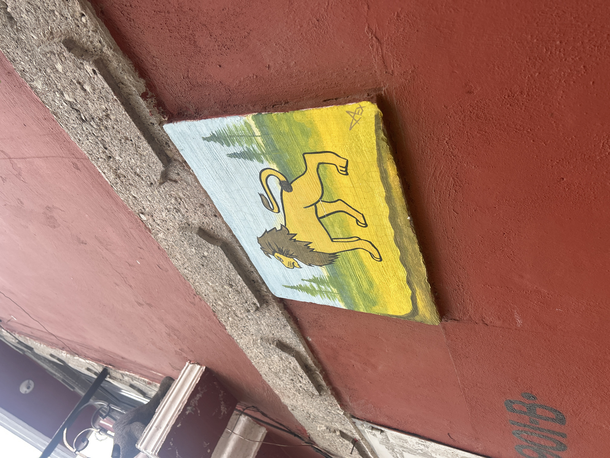 An outdoor, low-angle, medium shot captures a bright red building facade adorned with a painted tile depicting a yellow lion against a yellow and light blue background, with stylized green trees in the distance. The painting is positioned below a rough, textured concrete beam that runs horizontally across the image. To the left of the painting, a portion of another artwork or sign, predominantly white with black lettering, is visible, with the visible letters appearing to be "01B". The building's textured red wall exhibits minor imperfections. The scene suggests an urban environment, possibly in the city of Atlixco, Mexico, known for its vibrant colors and street art.