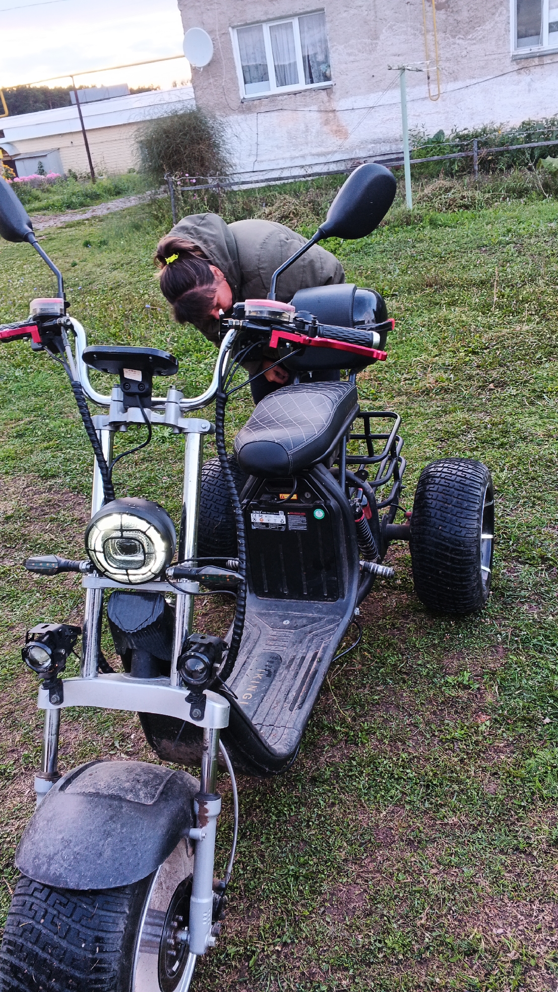 A person is leaning over a black electric scooter on a grassy area in front of a building in the village of Podgornoye, Russia. The scooter has three wheels, a padded seat, and a large circular headlight. The person is wearing a green hooded jacket and has their hair tied back with a yellow scrunchie. The building in the background is a light-colored, two-story structure with a satellite dish on the roof and several windows. There is a clothesline strung between two poles in front of the building. The ground is covered in grass and some bare patches of dirt. The sky is overcast.