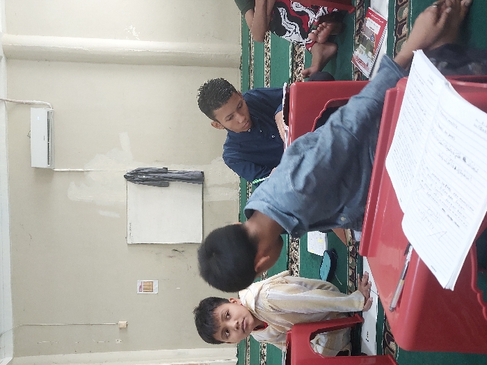 The image depicts an indoor scene in Lhokseumawe, Indonesia, likely a classroom or study area. The room features light-colored walls, displaying variations in paint tone and minor scuff marks, with a white split-unit air conditioner mounted high on the back wall. A white rectangular board, possibly a whiteboard, is mounted below the air conditioner to the left, with a dark garment hanging from a hook beside it. The floor is covered with a green carpet featuring a repeating patterned design in lighter green and red.
Four individuals, all male and appearing to be school-aged children or teenagers, are present. In the foreground, a boy wearing a denim-colored long-sleeved shirt is seated on the carpet, leaning forward. A red plastic stool serves as a low desk in front of him, holding an open notebook with visible writing and a pen. His left arm extends out of frame to the right. To his left, another boy in a striped light-colored long-sleeved shirt is seated on the carpet, looking towards the camera with a direct gaze. A red plastic stool is positioned in front of him. In the midground, to the right, a third boy with short dark hair, wearing a dark blue long-sleeved shirt, is intently bent over an open notebook resting on another red plastic stool, appearing to be writing or studying. To the far right, the bare feet of a fourth individual, seated on the carpet and partially obscured by a red plastic stool, are visible next to a red-covered book. A small blue pencil case is on the carpet near the boy in the striped shirt.
