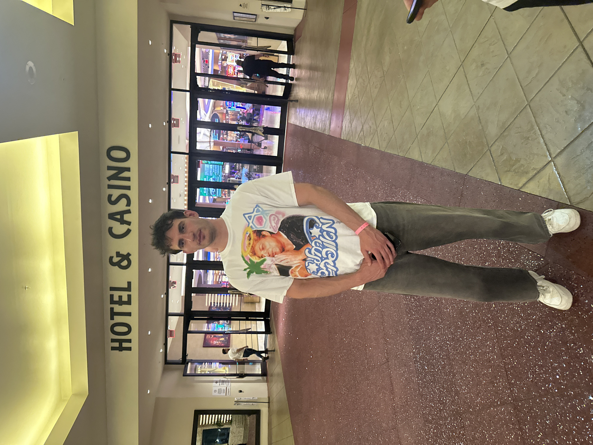 A man stands inside the Seminole Hard Rock Hotel & Casino in Hollywood Seminole Reservation, United States of America. He is wearing a white t-shirt with a graphic print, gray pants, and white sneakers. He has short dark hair. The casino entrance is visible behind him, with glass doors and people walking through. The interior flooring is a speckled reddish-brown. A sign reading "HOTEL & CASINO" is visible on a wall to the left. The lighting is bright and artificial.