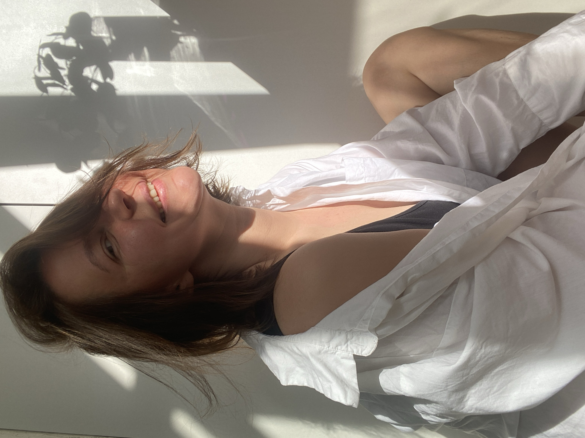A woman lies on a white surface in a brightly lit room. She wears a black top and a loosely draped white button-down shirt. Her head is tilted back, and she smiles, exposing her teeth. The scene is illuminated by natural sunlight, which creates sharp shadows on the white surface, including the shadow of a plant. The location is not discernible from the image, and the city of Chaikovsky, Russia, cannot be confirmed.