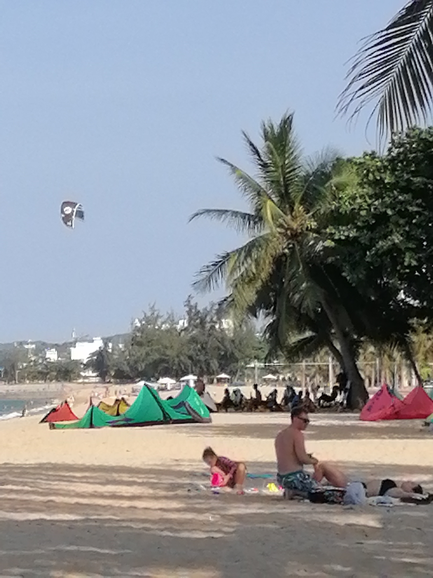 The image captures a sunny beach scene in Nha Trang Ward, Vietnam. A sandy beach slopes towards the ocean, lined with various green and red kites that lie on the sand. A kite sails in the sky. Palm trees line the coast. Buildings can be seen in the background. In the foreground, a man is sitting on the sand, seemingly applying sunscreen to another person who is lying down. A child plays near them.