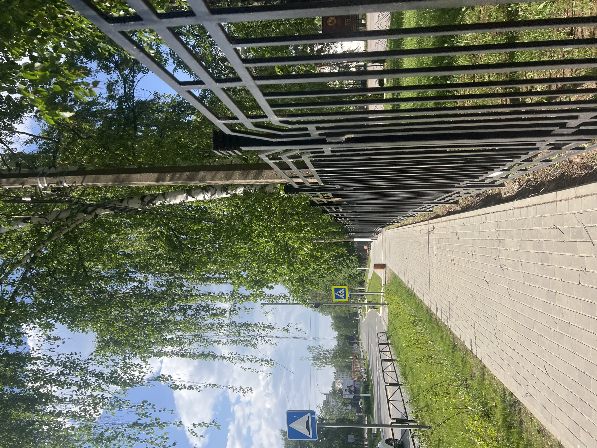 A street view in Chaikovsky, Russia, shows a paved sidewalk adjacent to a road, demarcated by a black metal fence. Green trees and a utility pole line the left side of the walkway, contrasting with the blue sky and scattered clouds visible overhead. Road signs indicate a pedestrian crossing and a bump or dip in the road. The fence appears to run parallel to the sidewalk, stretching into the distance. In the background, buildings and vegetation suggest a residential or suburban area.