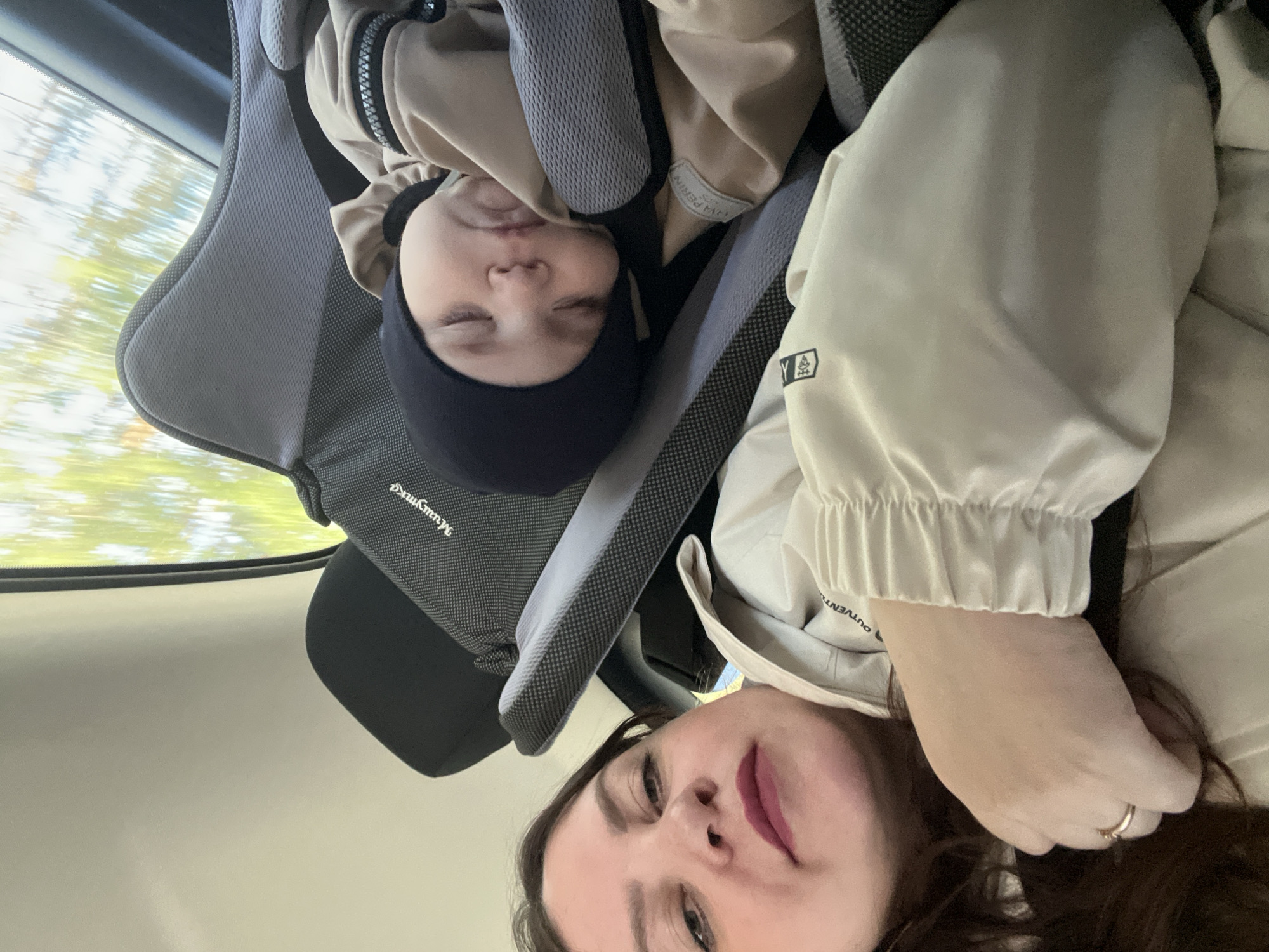 This medium-close-up image is taken inside a vehicle, likely a car, during daytime. The photograph is oriented at an angle, showing the subjects tilted. The primary subjects are an adult woman and a young child.
The child, appearing to be a baby or toddler, is positioned in a grey car seat, seemingly asleep or resting peacefully with eyes closed. They are wearing a dark beanie and a beige jacket. Text "Mummykid" is visible on the car seat's fabric, and part of a label reading "KAYA PEREIRA" is seen on the child's jacket.
The adult woman occupies the foreground, looking towards the camera with a neutral to slightly smiling expression, wearing pinkish-red lipstick. She has dark hair and is dressed in a light beige or off-white jacket. Her left hand is visible near her shoulder, adorned with a simple gold ring on her ring finger. Text "OUTVENTURE" is visible on the cuff of her jacket sleeve.
Through the car window in the upper left corner, blurred green and yellowish hues suggest trees or foliage, indicating that the vehicle is in motion. The natural light filtering in confirms it is daytime. The scene conveys a moment of travel or a road trip. The provided location context for the image is Ol'khovochka, Russia.