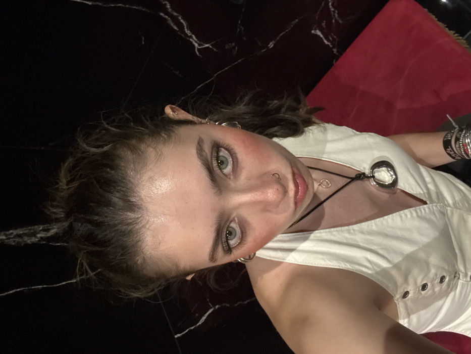 A woman with light skin, gray eyes, and rosy cheeks looks up at the camera. She wears a white top with metal grommets and a black choker necklace. A large silver pendant hangs from the necklace. She has multiple silver hoop earrings. The background includes black marble flooring with white veining and a red cushion.