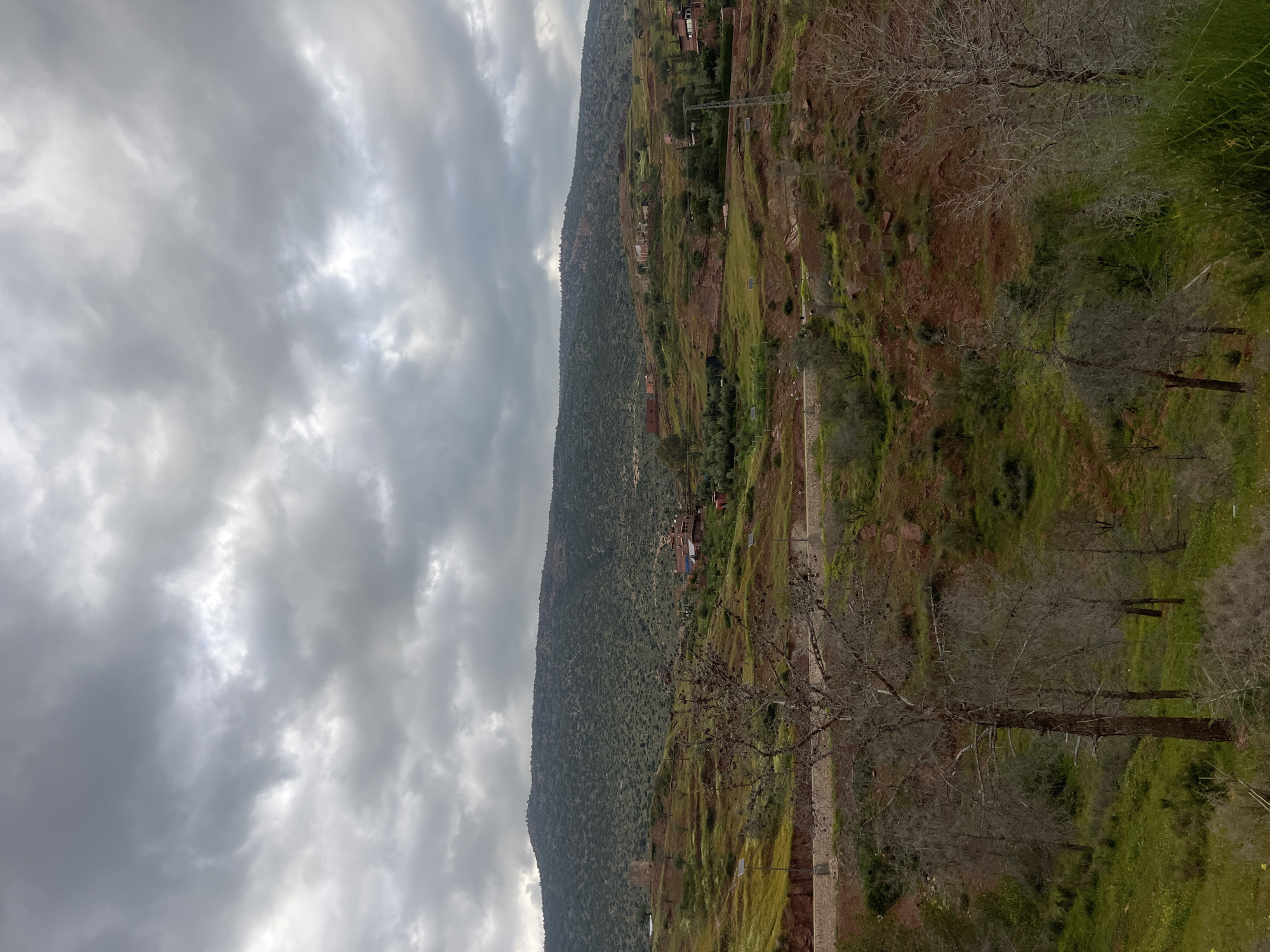 The image depicts a landscape near Bin El Ouidane, Morocco, under a cloudy sky. A steep, terraced hillside dominates the scene, displaying a mix of green vegetation and red soil. Scattered buildings and structures are visible along the slope. In the foreground, bare trees and green foliage provide a closer view of the environment. A stone or concrete pathway runs along the hillside. The overall impression is one of a rural, mountainous area with evidence of human habitation and cultivation.