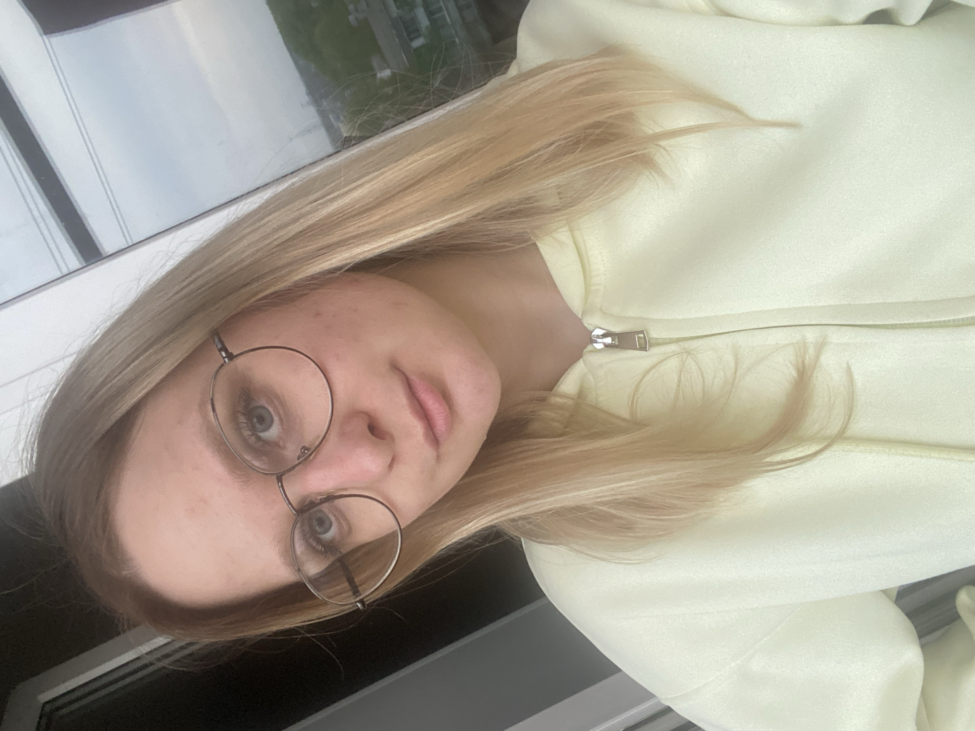 A fair-skinned young woman with light blonde hair is seen in a close-up selfie. She is wearing round, wire-rimmed glasses and a light yellow zip-up jacket. The background shows an out-of-focus view of buildings, suggesting an urban setting. Based on the user's specification, the location is identified as Chaikovsky, Russia. The specific location within Chaikovsky is indeterminable from the image.