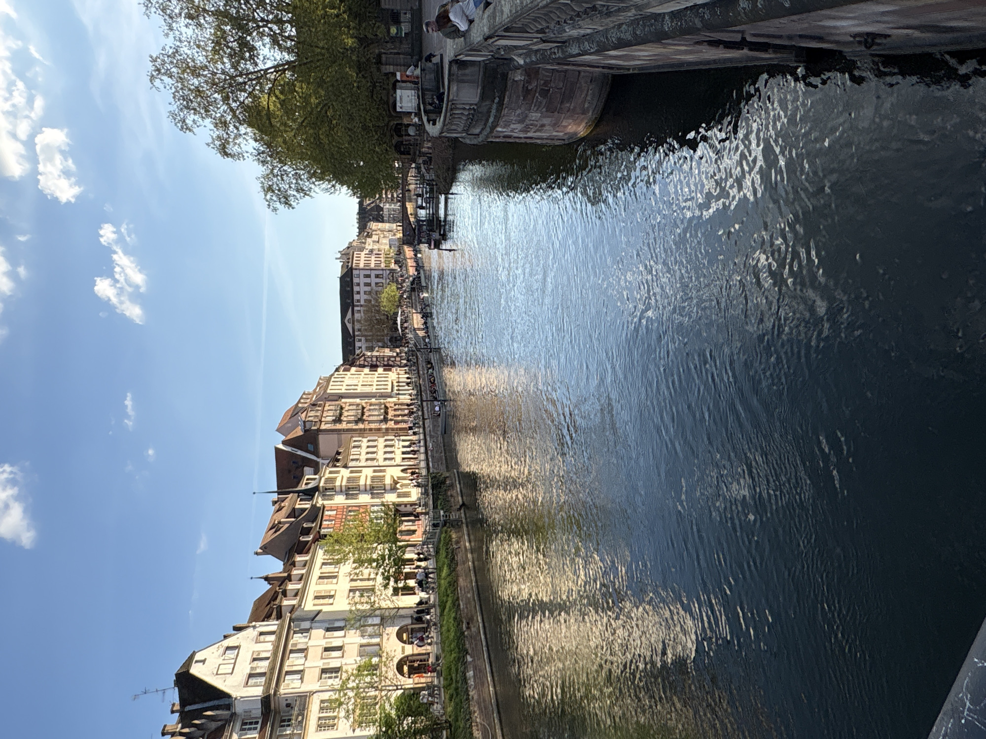 The image shows a canal in Strasbourg, France, likely the Ill River, lined with traditional Alsatian buildings. The buildings are characterized by half-timbered facades and pitched roofs. Along the canal's edge, people are sitting and walking, enjoying the waterfront. A bridge crosses the canal in the foreground. The water is dark with reflections of the buildings and sky. The sky is blue with scattered clouds. A tree is visible on the left, partly obscuring the buildings.
