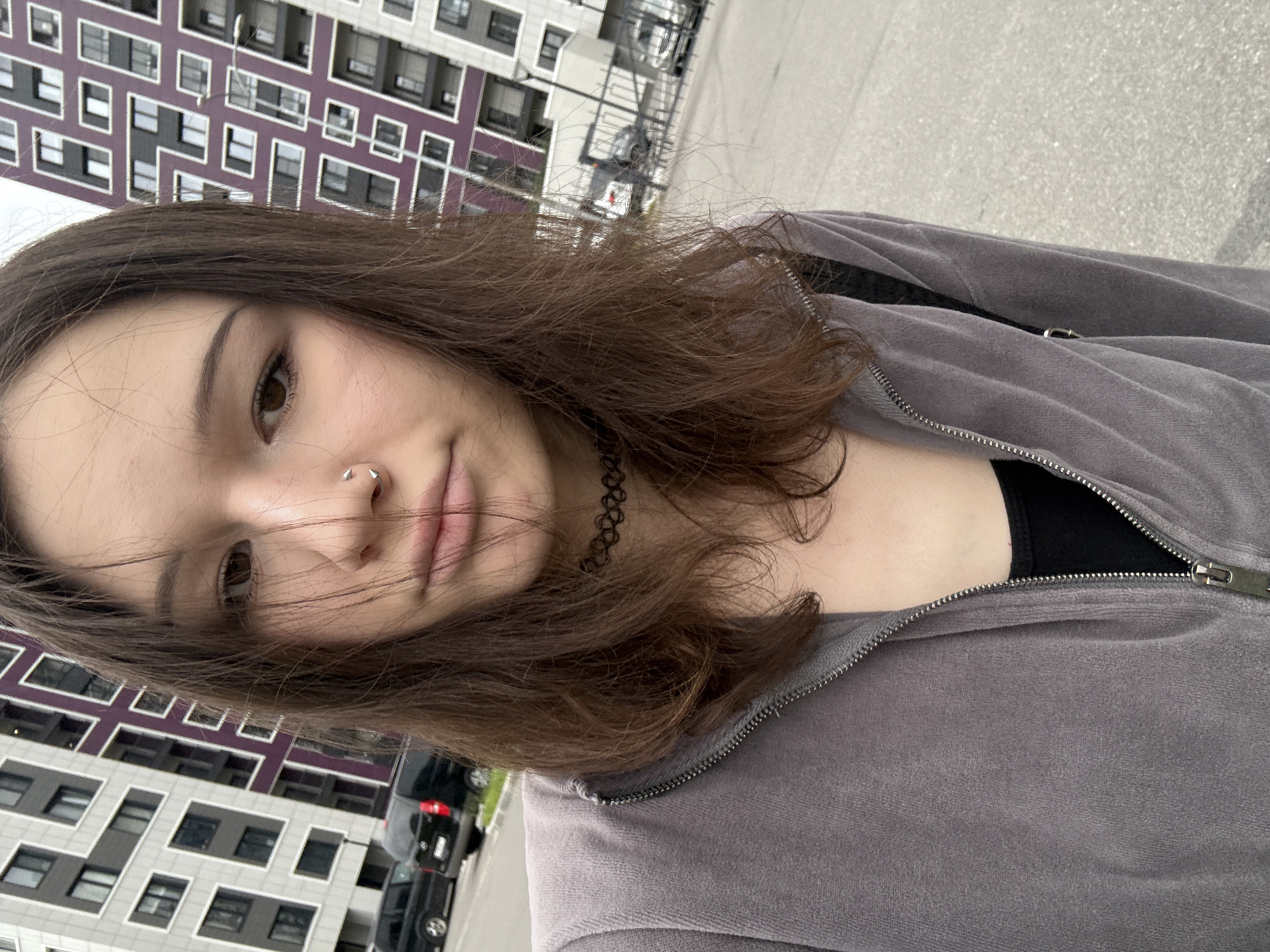 A young woman with dark brown hair is taking a selfie in front of a modern apartment building. She is wearing a grey zippered hoodie over a black tank top and a black choker necklace. She has a nose ring. The apartment building has a geometric design with purple and grey accents. In the background, there are cars parked and a fence. The ground appears to be asphalt. This location is in the city of Kokkorevo, Russia.