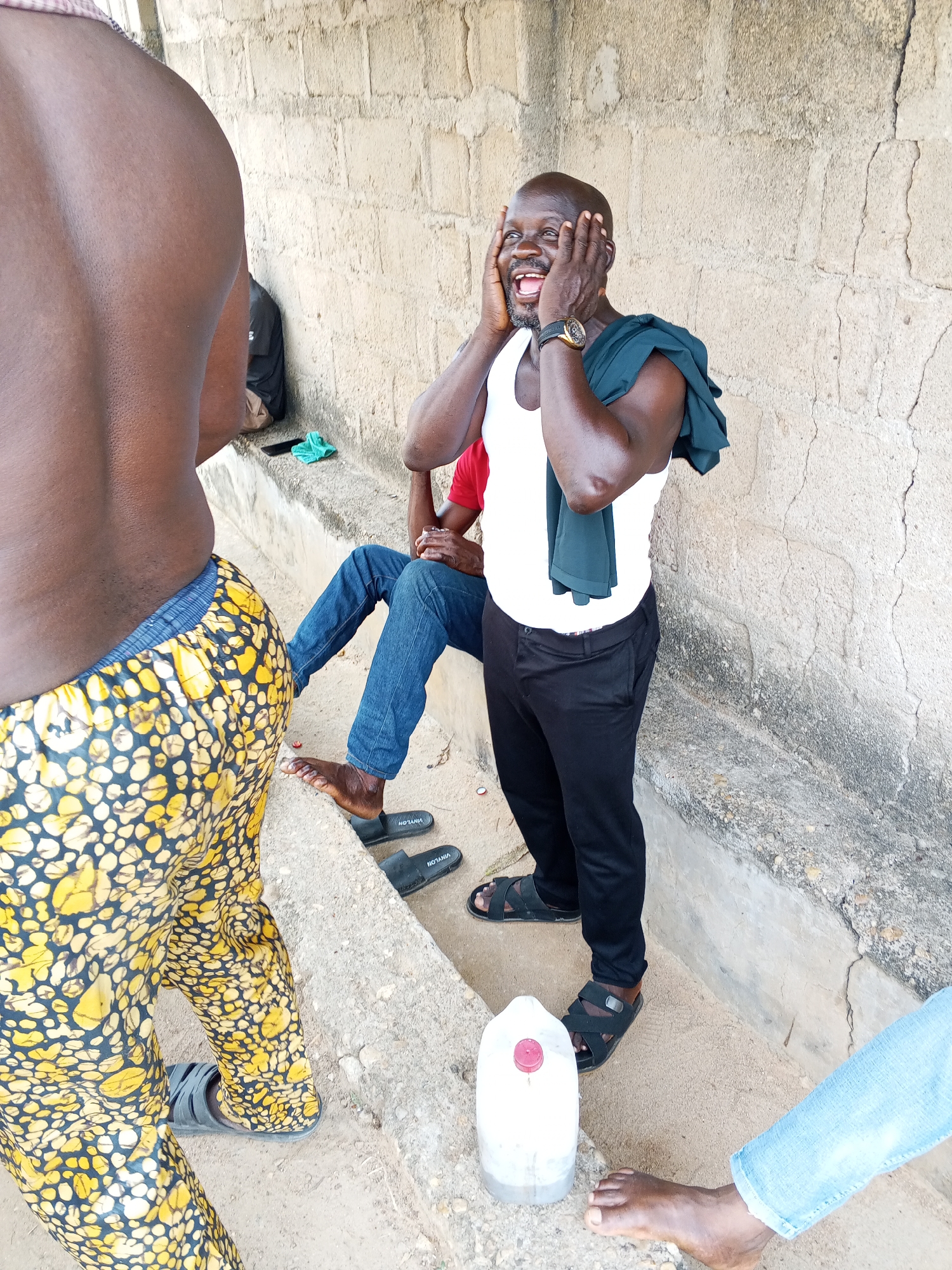 The image depicts a candid scene in Adonkolo, Nigeria. A group of people are gathered outdoors near a weathered, light-colored concrete structure. One man, wearing a white tank top and black pants, is laughing with his hands on his face, a dark green garment draped over his shoulder. Another individual, seen from the back, is wearing patterned yellow and black pants. A person in blue jeans is seated on a concrete ledge, and a plastic container with a red cap sits on the ground. There is another man wearing jeans with a red t-shirt. Several pairs of sandals are scattered on the ground. The setting appears to be casual and relaxed.