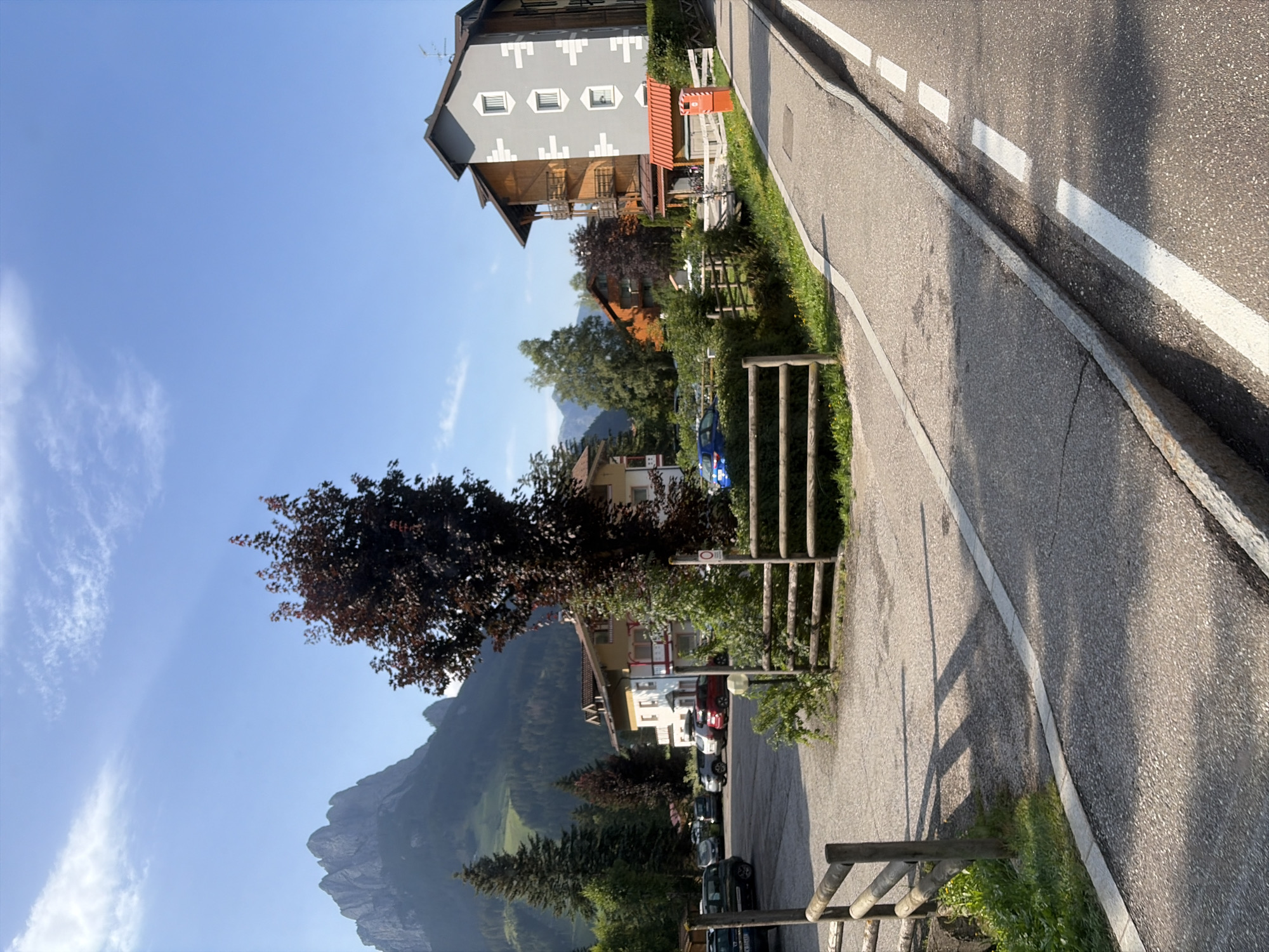 This is a sunny, outdoor, eye-level, medium shot of a street scene in Pera, Italy. The scene features a parking lot with several cars, a wooden fence, a sidewalk, and a road. In the background, there are buildings, trees, and mountains. The sky is blue with some clouds. The overall atmosphere is peaceful and serene.