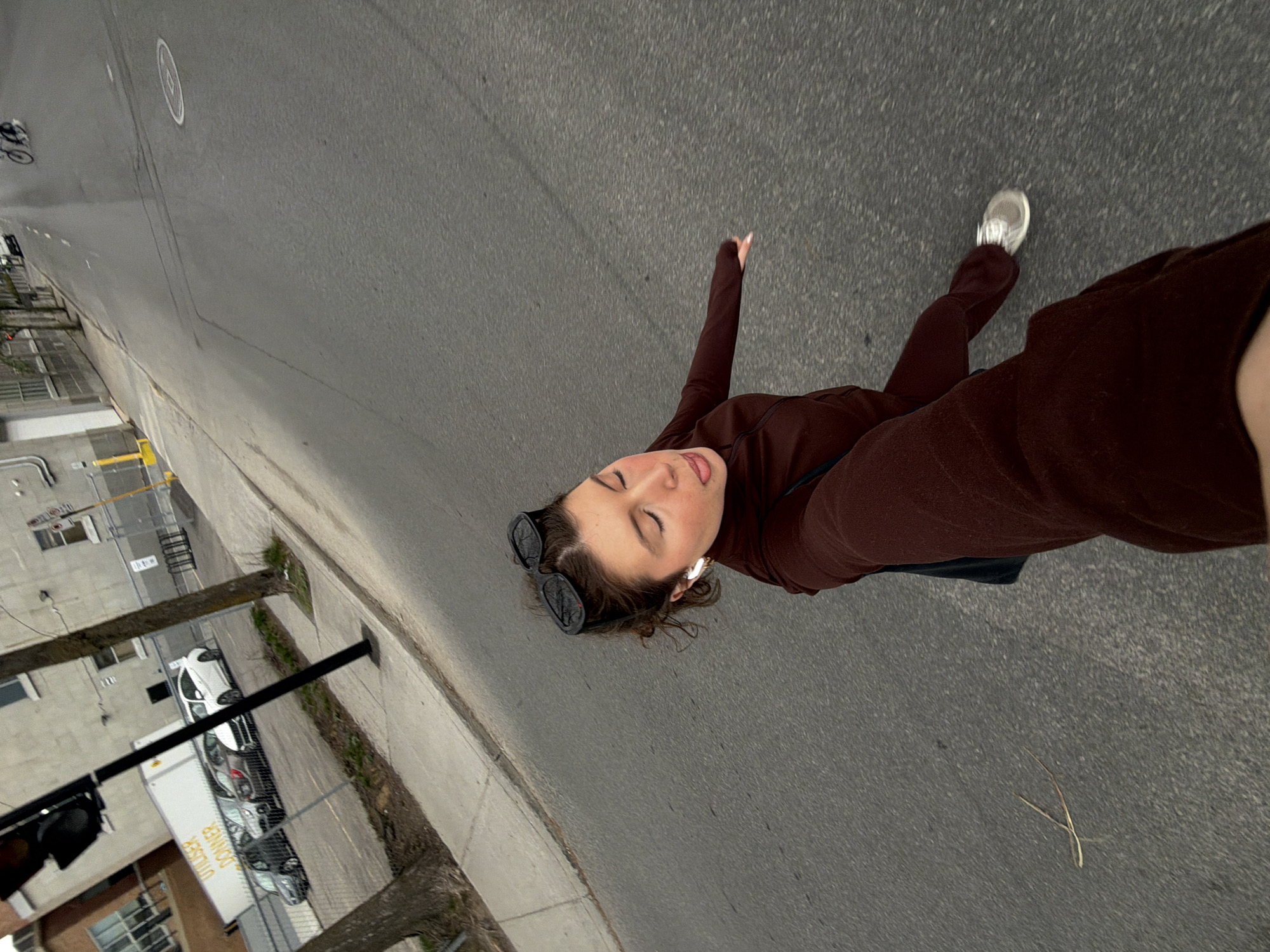 A young woman in a brown top and pants is walking on the sidewalk next to a street in Montreal, Canada. She is wearing sunglasses and an earpiece. The street is paved and there are buildings, trees, and parked vehicles on the other side of the sidewalk. A bicycle is visible in the distance.