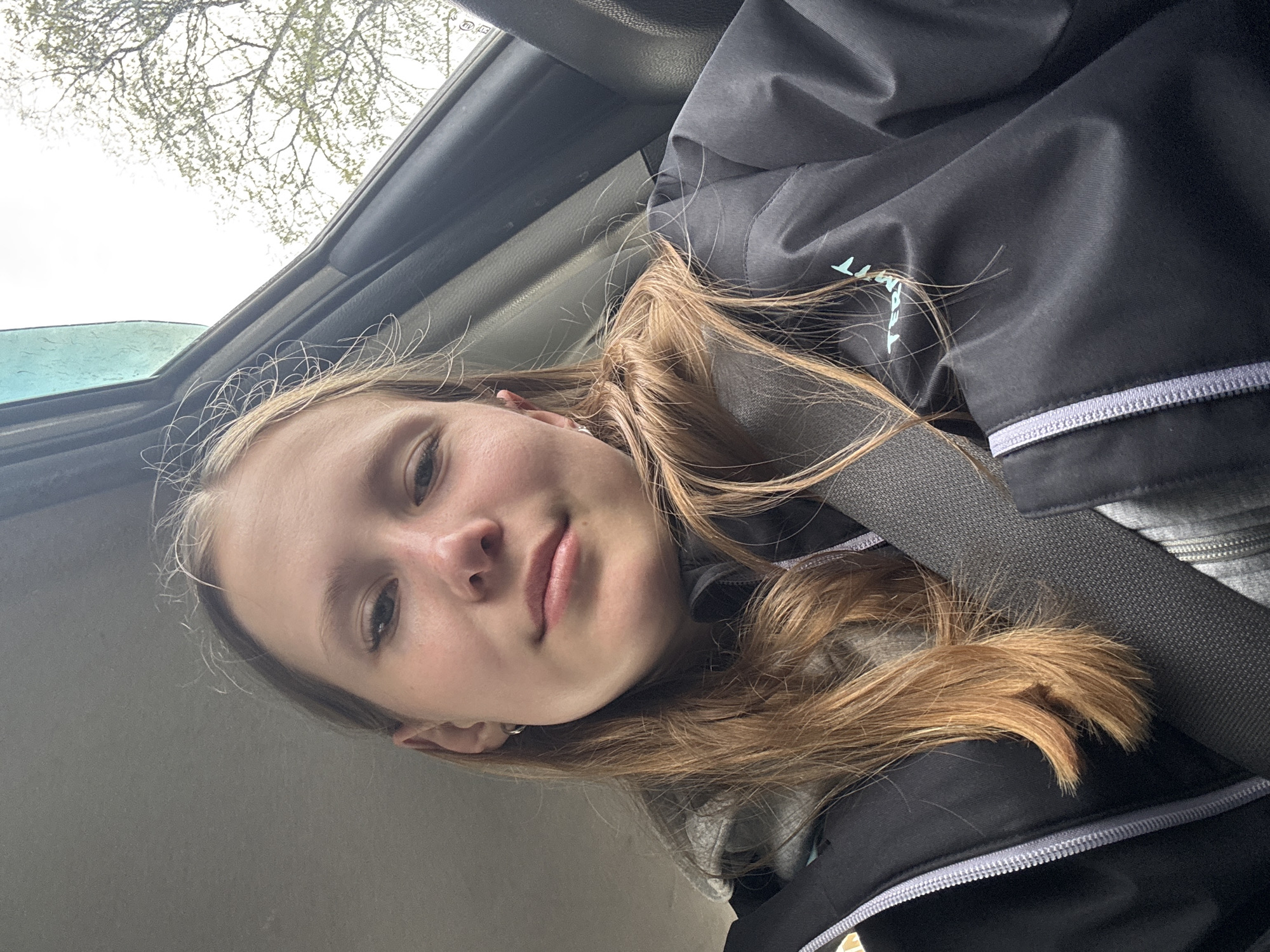 A young person with light brown hair is sitting in a car, wearing a seatbelt and a black jacket with a grey stripe at the cuff. They are looking directly at the camera with a slight smile. The sky is visible through the car window, along with some tree branches. The location cannot be determined from the image alone.