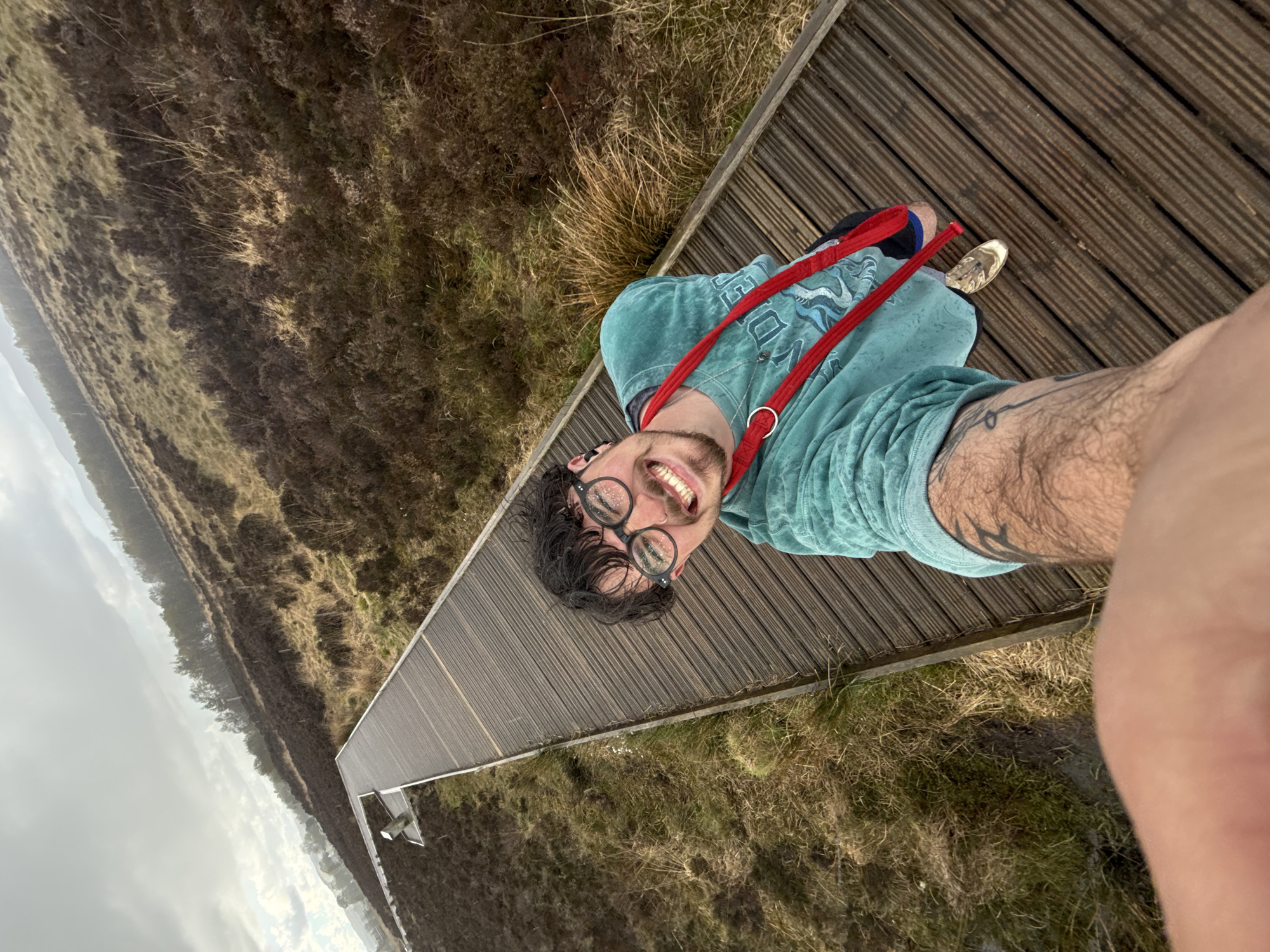 A man is taking a selfie while standing on a wooden boardwalk in a grassy, field-like area. The boardwalk extends into the distance. He is wearing glasses, a blue-green shirt, and a red strap across his chest. A glimpse of his shoe can be seen, and there is a tattoo on his arm. The sky is overcast. The location appears to be a natural area with low-lying vegetation.