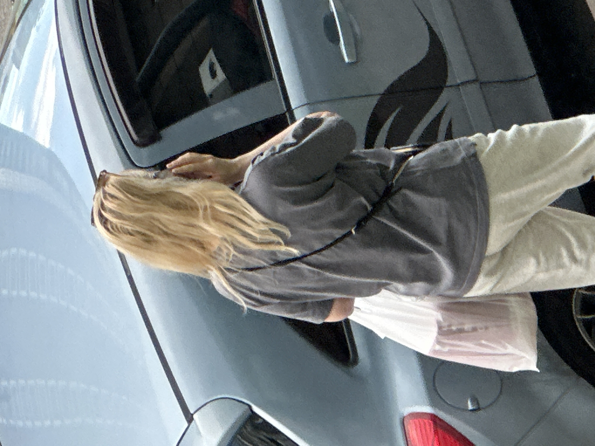 A woman with blonde hair is standing next to a light blue car. She is wearing a gray t-shirt and light-colored pants. She is holding a white plastic bag in her left hand and appears to be talking on her phone with her right hand. The car has a black graphic design on its side. The background is blurred, but appears to be an outdoor urban environment. The image was taken in Murino, Russia.