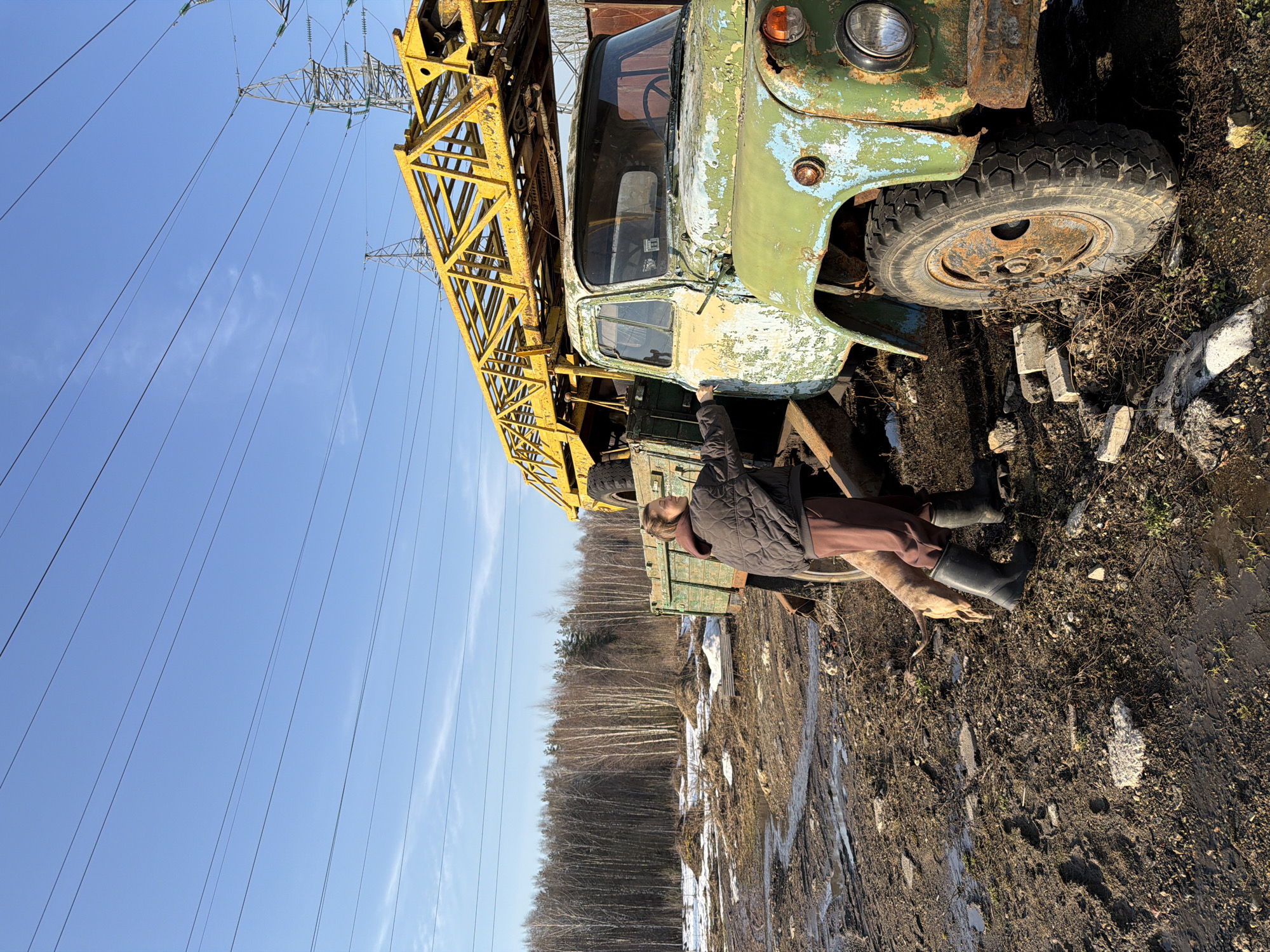 This image depicts an outdoor scene in Khimki, Russia, under a clear blue sky. Multiple high-voltage power lines span the upper frame, supported by at least two metal lattice transmission towers in the background.
A single individual stands alongside a severely dilapidated, older model truck in the mid-ground. The truck exhibits extensive rust and peeling paint, with original colors appearing as faded green and light blue. A large, yellow, corroded lattice-work structure, possibly part of a crane or drilling rig, is mounted on its bed, extending significantly above the cab. The person, appearing female, wears a dark grey quilted jacket, brown pants, and dark rubber boots. Her left hand is positioned near the driver's side door frame of the truck. A light brown dog stands adjacent to the person's legs, partially obscured by her body.
The immediate ground is an uneven expanse of dark brown mud, interspersed with patches of melting snow, ice, and scattered concrete debris. Numerous footprints are visible in the mud. A dense line of bare deciduous trees forms the horizon on the left side of the frame.
