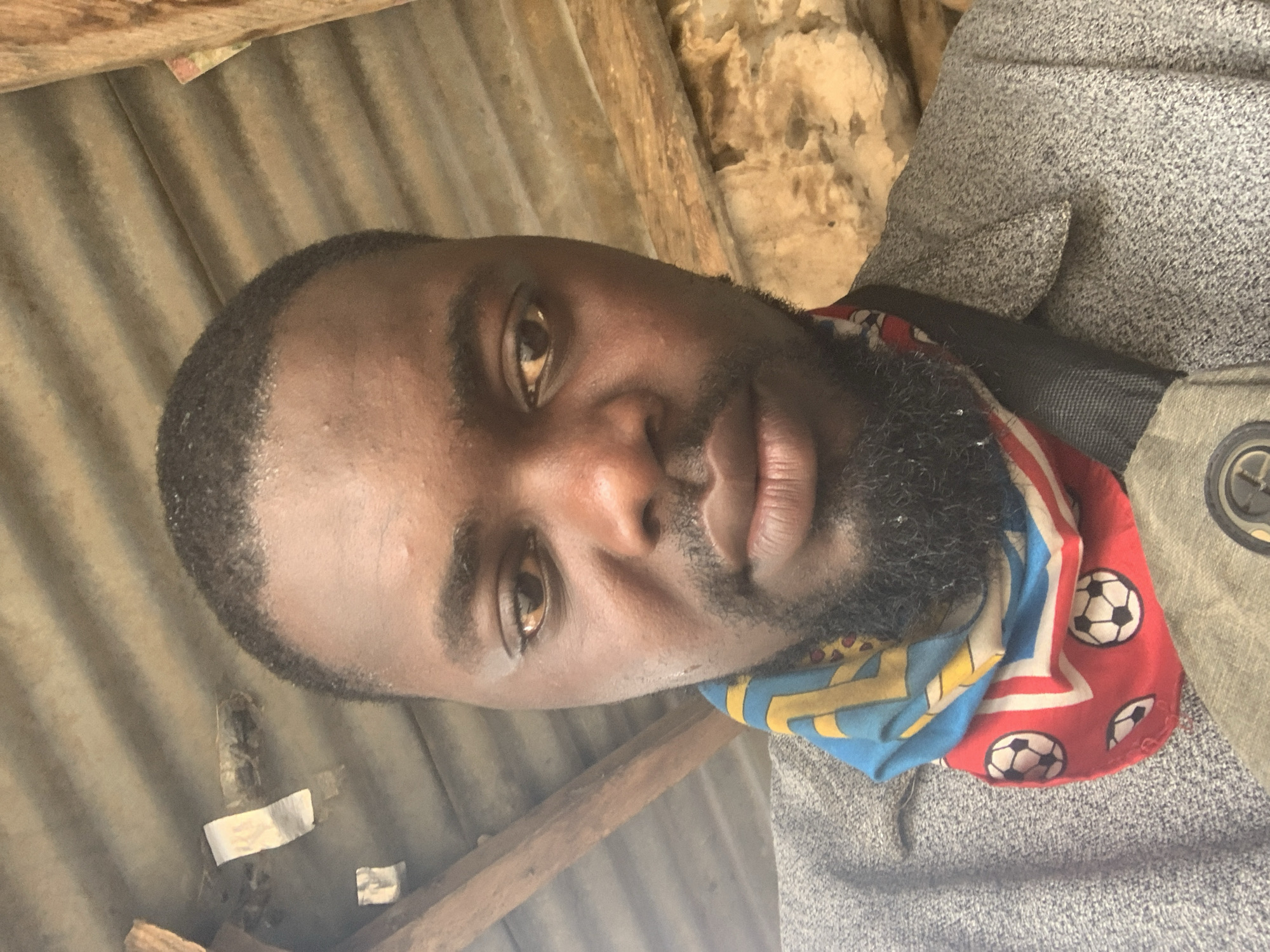 A dark-skinned man with a short beard and hair is captured in a close-up selfie. He is wearing a gray jacket and a colorful bandana around his neck featuring soccer balls and geometric patterns. The man's face is illuminated by natural light, and his expression is neutral. The background appears to be an outdoor setting with a corrugated metal roof and wooden beams overhead. Behind him, a rough, stone-like texture is visible. Given the context of Shagamu, Nigeria, this scene likely depicts a moment in a residential or semi-rural environment, possibly under a shelter or outbuilding.