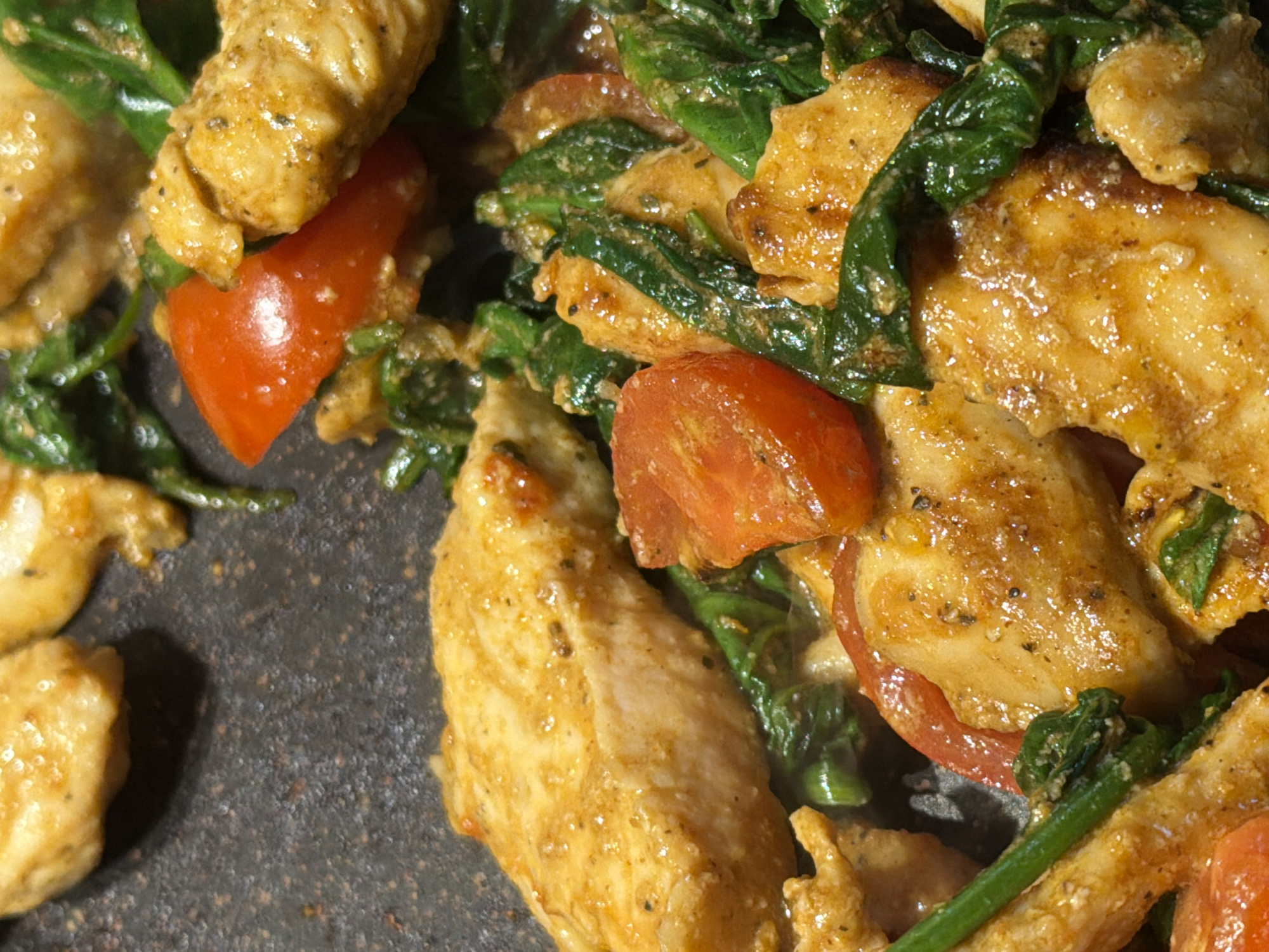 A close-up, overhead shot shows cooked chicken strips, cherry tomato halves, and wilted spinach in a pan. The chicken is golden brown and appears to be seasoned. The tomatoes are bright red and glistening. The spinach is dark green and appears to be cooked down. The pan is dark and textured, suggesting it is a cast iron skillet or similar cooking surface. The lighting is warm and highlights the food. There are no people or any specific location details visible in the image.
