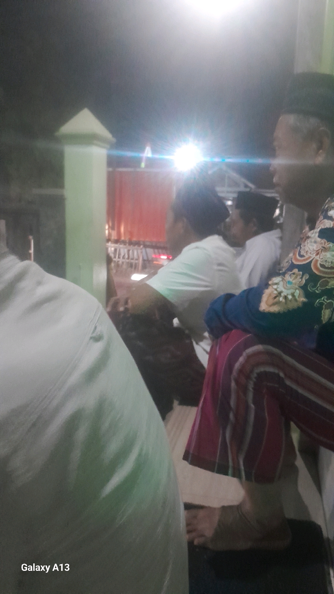 The image depicts a gathering of at least three individuals in what appears to be an outdoor or semi-outdoor setting at night. The lighting suggests artificial illumination, with bright lights causing lens flares. In the foreground, a large object, possibly a person in a white garment, is partially visible on the left. To the right, a man wearing a black peci (cap) and a patterned blue and gold garment is seated, with his profile visible. He is looking towards the right. Another man, wearing a white t-shirt and a darker garment, is seated to his left and slightly behind him. A fourth individual, also wearing a peci, is partially visible behind the man in the white t-shirt. The background includes a light-colored pillar on the left, a red structure in the middle ground which could be a tent or building, and rows of vertical elements suggesting a fence or scaffolding. The overall ambiance is subdued, likely due to the night time setting. The location is identified as Ngelo, Indonesia.