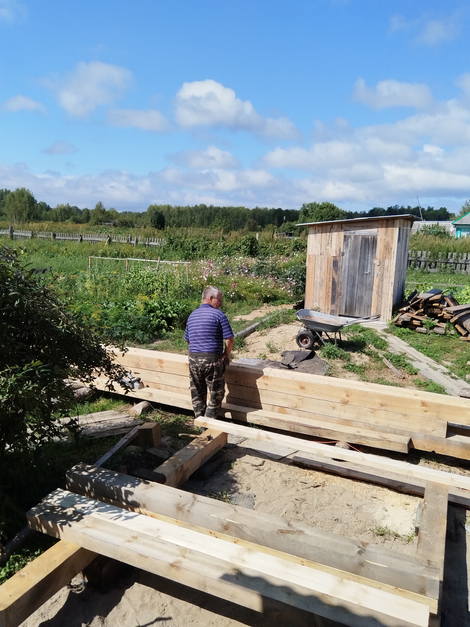 A man stands in an outdoor construction area in Minayevka, Russia, with a wooden shed in the background. He is wearing a striped shirt and camouflage pants. The ground is covered with lumber, some of which is being used to build a foundation. A wheelbarrow is visible near the shed, and a pile of cut wood is stacked to the right of it. The environment appears to be rural, with trees and a fence in the distance, under a bright blue sky with scattered clouds.