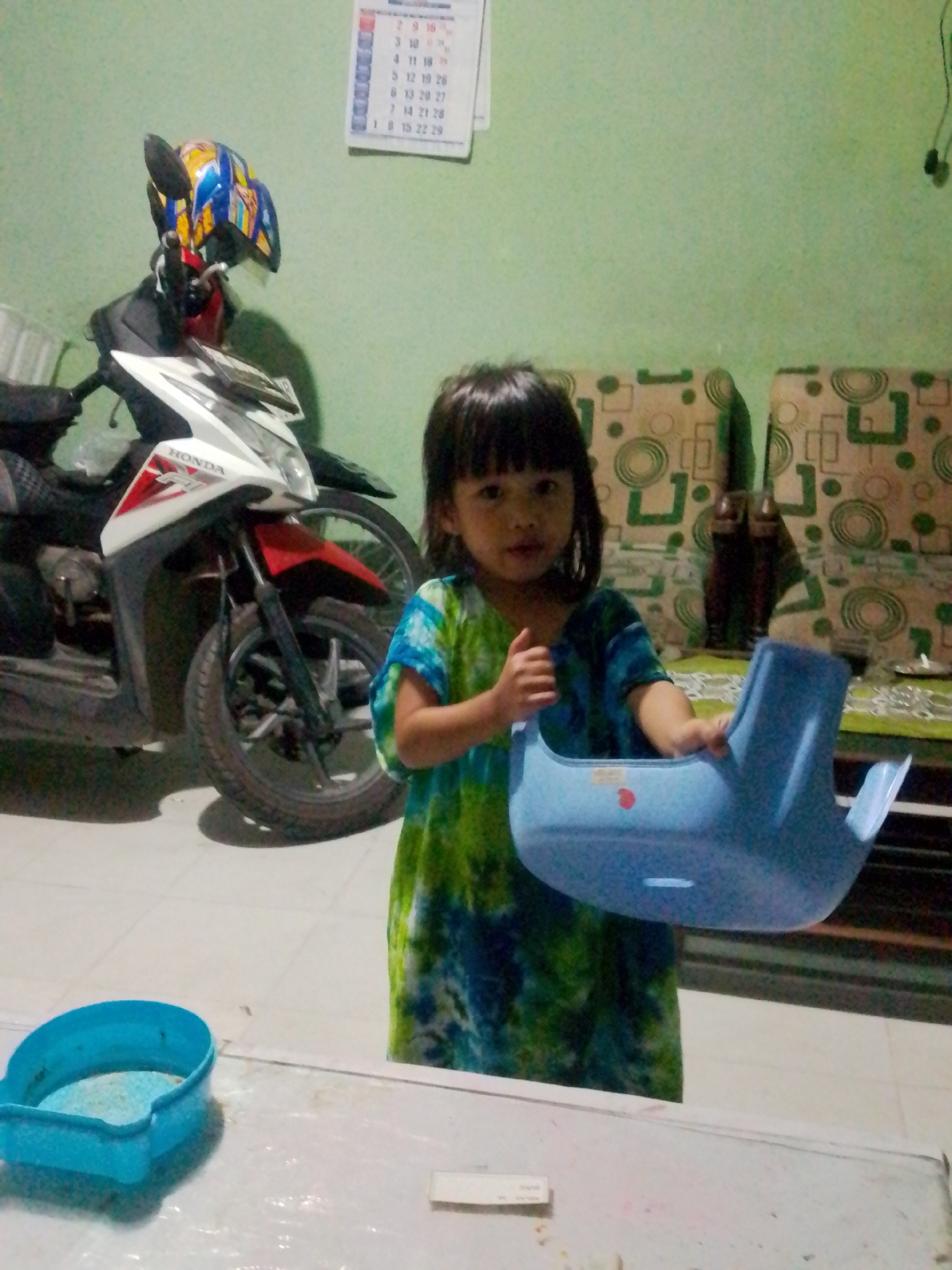 The image depicts an indoor scene in Bandar Lampung, Indonesia, likely a residential area. A young girl, approximately 4-6 years old, stands in the foreground. She has dark hair and is wearing a green and blue tie-dye dress. She is holding a blue plastic object, possibly a toy or part of an item, in her right hand and giving a thumbs-up with her left hand. To her left is a white and red Honda motorcycle with a blue, yellow, and red helmet placed on its handlebars. In the background, a green wall is visible, with a calendar hanging on it and a patterned sofa. A light blue plastic container with some white material inside sits on a white surface in the bottom left corner of the frame. The lighting suggests it is nighttime or indoors with artificial lighting.