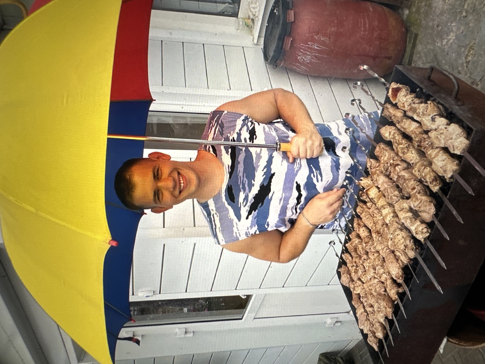 The image depicts a young man standing under a colorful umbrella, holding it with his left hand. He is wearing a sleeveless shirt with a camouflage pattern in shades of blue and white. The man is smiling and looking towards the camera. In front of him is a charcoal grill filled with numerous skewers of raw meat, suggesting he is preparing to cook them. Behind the man is a white wooden building with horizontal siding. To the right of the grill, a large red barrel is visible. The scene appears to be an outdoor setting, possibly a backyard or a picnic area. The lighting suggests it is daytime. The specific location in Tallinn, Estonia cannot be determined from the visual information provided.
