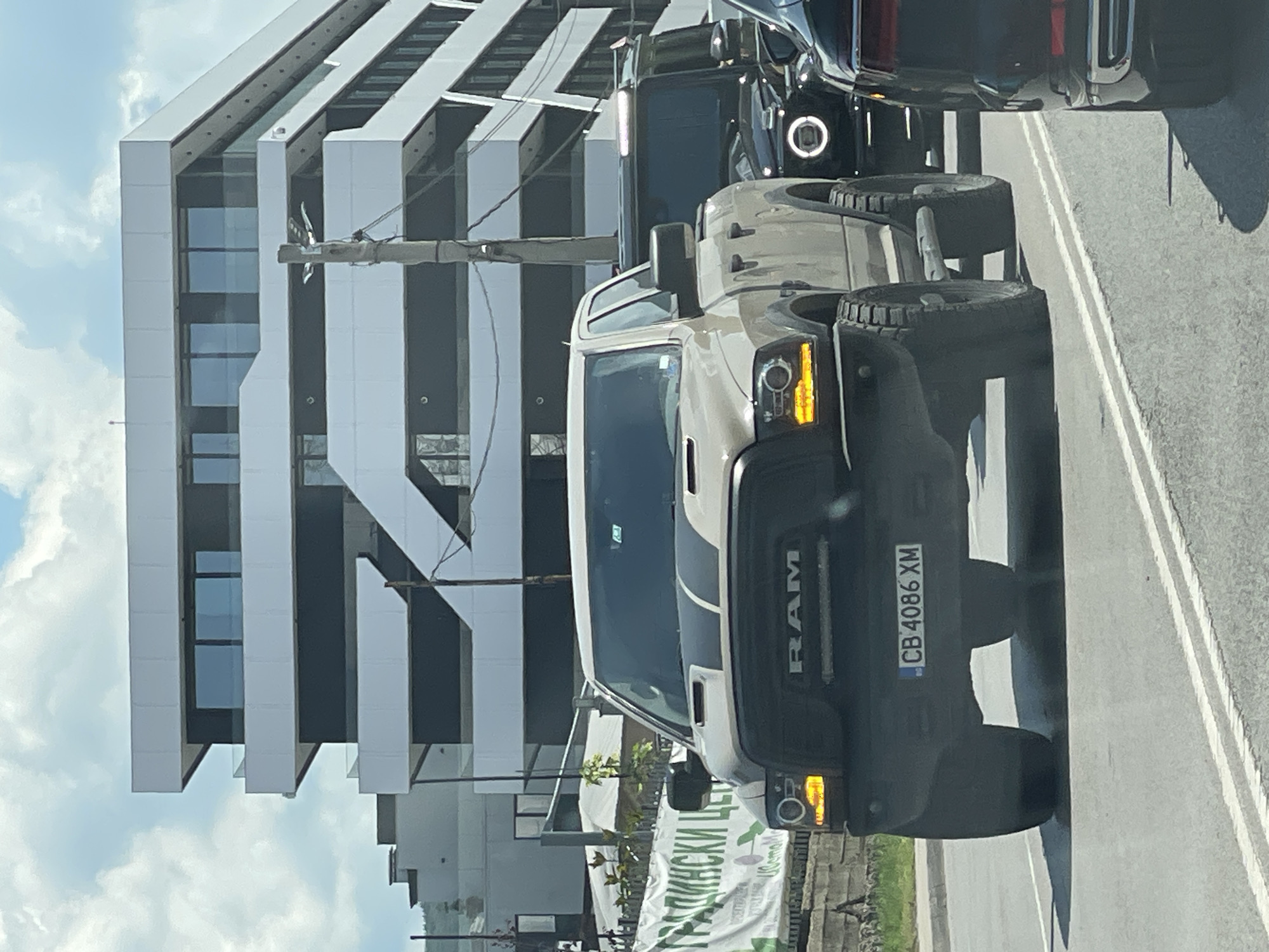 The image captures a traffic scene in Sofia, Bulgaria, on a cloudy day. In the foreground, a tan RAM pickup truck is prominently featured, with its headlights illuminated and a black stripe running down its hood. The truck's license plate reads "CB 4086 XM." Behind the RAM truck, a black SUV, possibly a Mercedes-Benz G-Class, is visible with its distinctive circular headlights on. To the far right, the rear of a dark-colored vehicle is partially visible. In the background, a modern building with a striking geometric design, featuring white facades and dark-tinted windows, dominates the skyline. A utility pole with wires stands between the traffic and the building. A white banner with Cyrillic text, appearing to advertise a garden center, is visible on the left side of the image, near the curb. The vehicles are on a paved road with white lane markings, indicating they are in traffic.