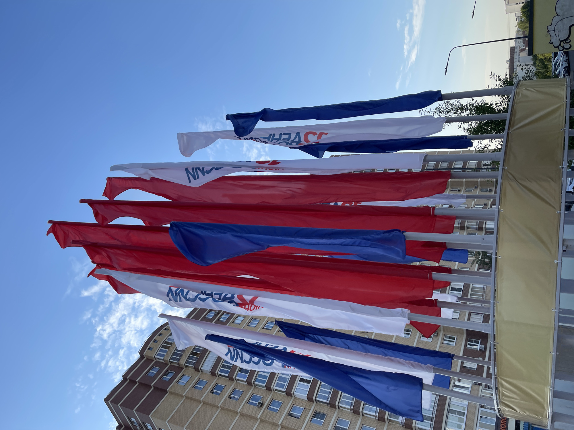 The image shows a cluster of red, white, and blue flags on poles, set against the backdrop of a modern apartment building and a clear blue sky with wispy clouds. The flags are predominantly red and white, with some featuring blue sections, possibly representing the Russian flag. Several flags have text and logos printed on them, some of which are partially obscured. The building appears to be a residential high-rise with numerous windows. In the foreground, a golden banner is visible, stretched around the base of the flagpoles. The scene suggests an outdoor public space, likely in a city, given the urban architecture and the presence of streetlights in the background. The lighting indicates it is daytime, with the sun casting shadows.