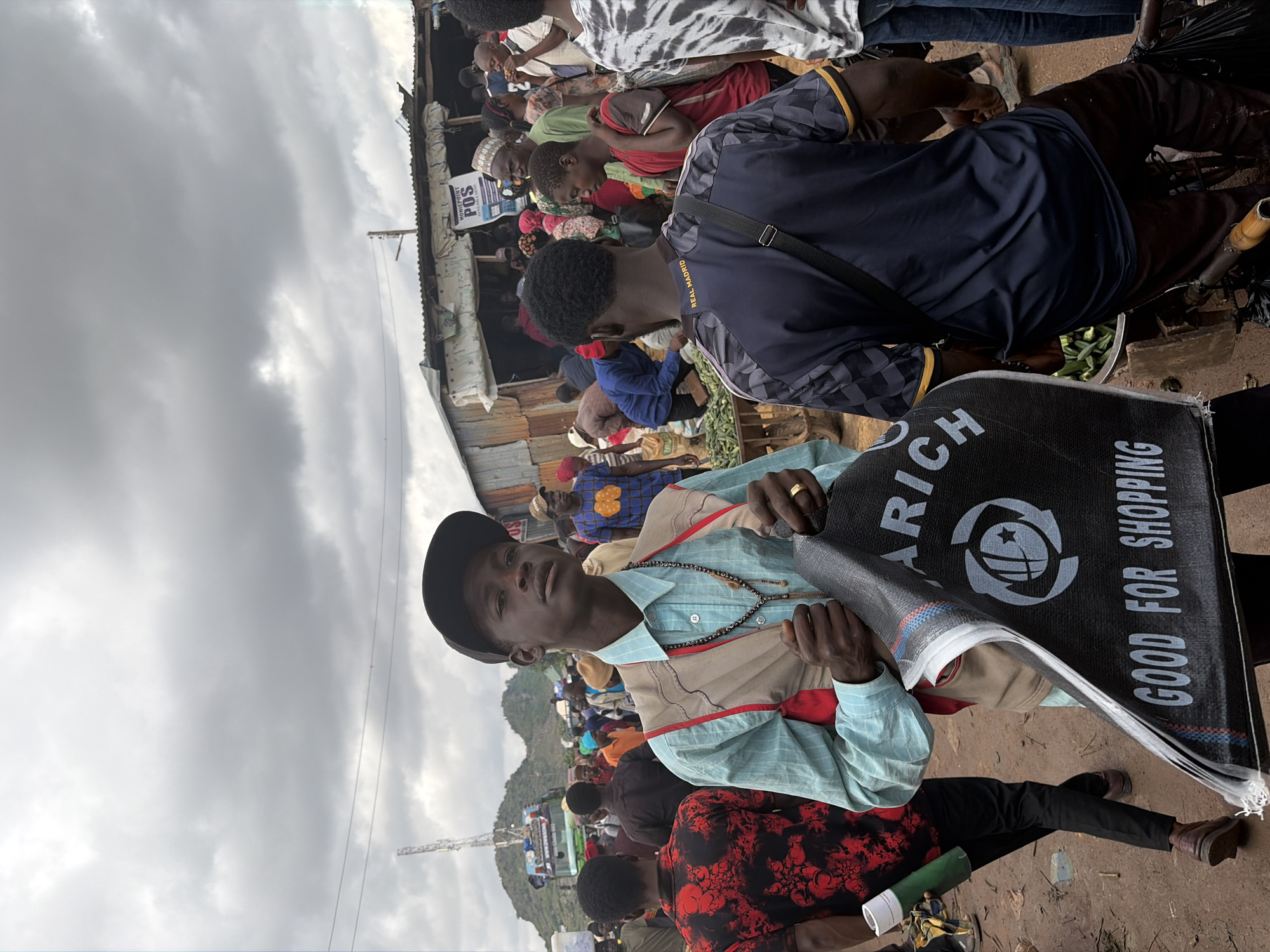 The image depicts a bustling open-air market in Gura Top, Nigeria. The sky is overcast with thick, gray clouds. In the foreground, a young man wearing a light blue collared shirt, a tan vest, and a black baseball cap holds up a large, black shopping bag with white lettering that reads "ARICH GOOD FOR SHOPPING". He wears a necklace and a ring on his left hand. Behind him, other market-goers are visible, some carrying goods and others browsing stalls. One man in the mid-ground wears a dark blue shirt with yellow accents and a black shoulder strap. In the background, a line of makeshift stalls constructed from corrugated metal sheets are set up. A sign indicating "MONIEPOINT POS AVAILABLE HERE" is visible on one of the stalls. A tower structure and a truck with a green and white body are seen in the distance, suggesting a roadside market. The ground is dirt, and the overall impression is one of lively commerce in a somewhat rudimentary setting.
