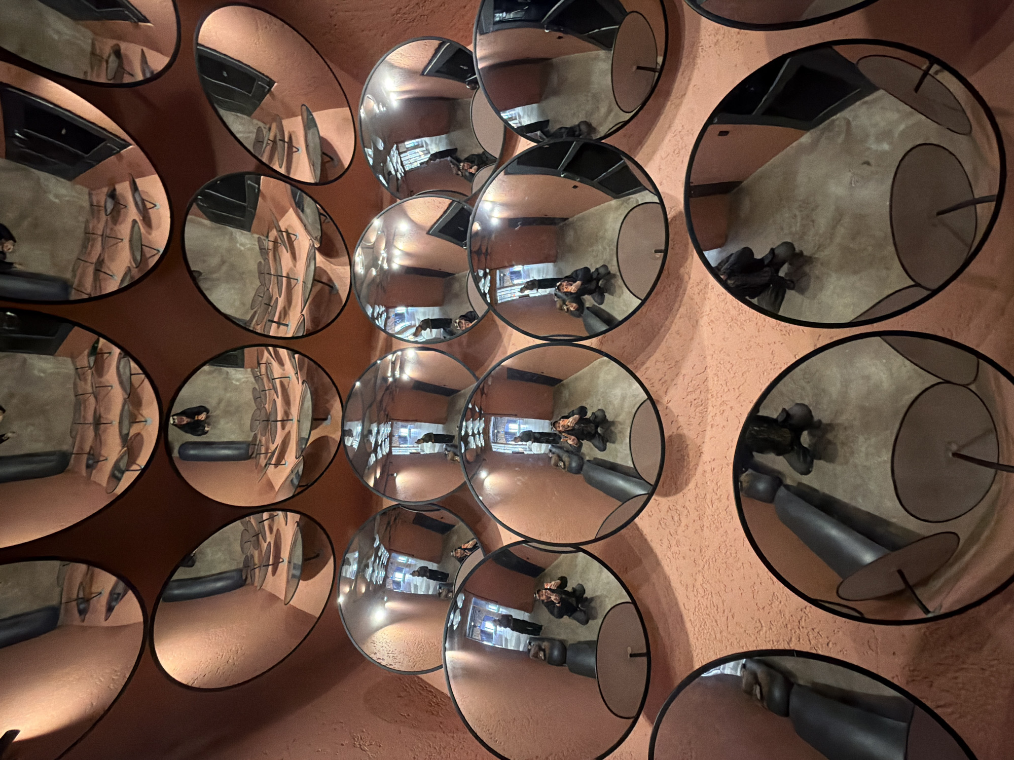 The image shows a wall in what appears to be a room, covered with circular mirrors. The mirrors reflect the interior of the room, including people, furniture, and architectural details. The room itself is painted in a warm, reddish-brown color. The reflections within the mirrors distort the scene, creating a disorienting effect. Based on the objects reflected in the mirrors, including the mannequins and furniture, it suggests a fitting room in a clothing store. The scene suggests an art installation or a boutique with a unique design. The location is likely Saint Petersburg, Russia.