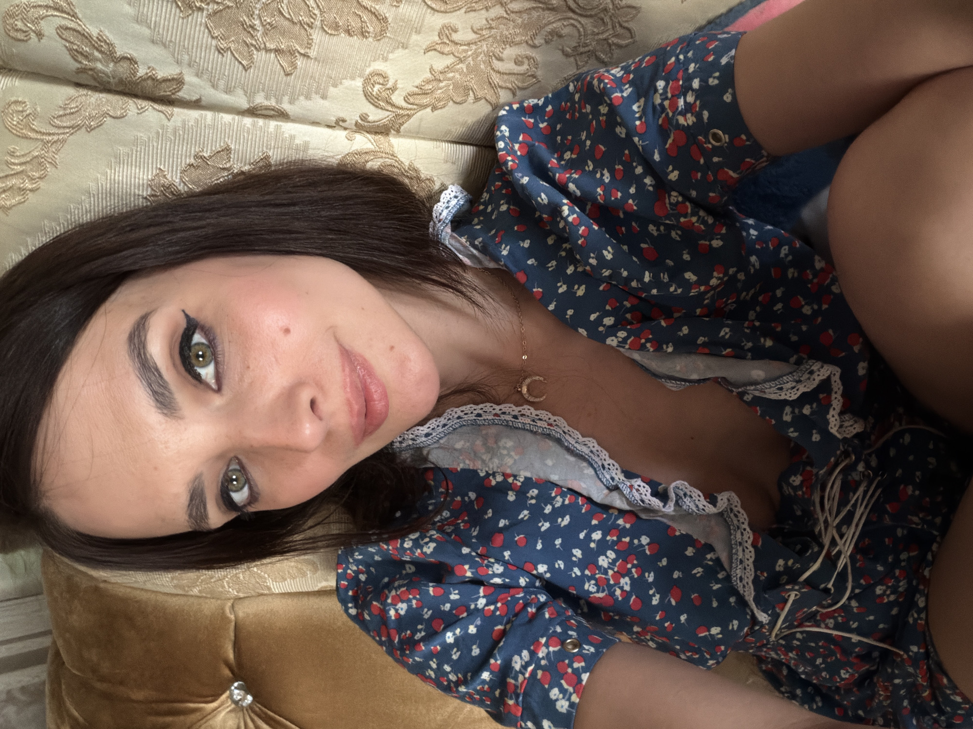 A close-up, overhead shot features a young woman with dark hair and green eyes lying on a textured, gold-colored sofa. She is wearing a dark blue dress with a pattern of small red and white flowers. The dress has white lace trim around the neckline and sleeves, and a lace-up detail on the bodice. A delicate gold necklace with a crescent moon pendant is visible around her neck. Her makeup is subtly applied, with winged eyeliner and rosy lipstick. The lighting suggests it is daytime, possibly indoors, given the soft light and the presence of a sofa. The overall mood appears relaxed and intimate. There is no visible text in the image. The provided context indicates the location is Ufa, Russia.