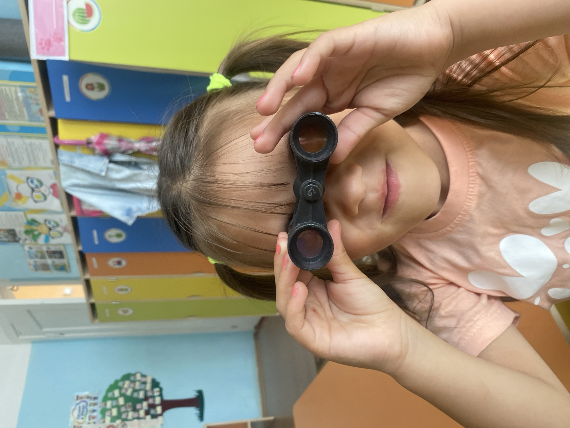 A young girl with pigtails and a pink shirt is looking through black binoculars. She is holding the binoculars up to her eyes with her hands. The background appears to be a classroom or daycare with colorful lockers and decorations on the walls. The overall setting suggests an indoor educational environment. There are no discernible landmarks or clues that would indicate the specific location in Chaikovsky, Russia.
