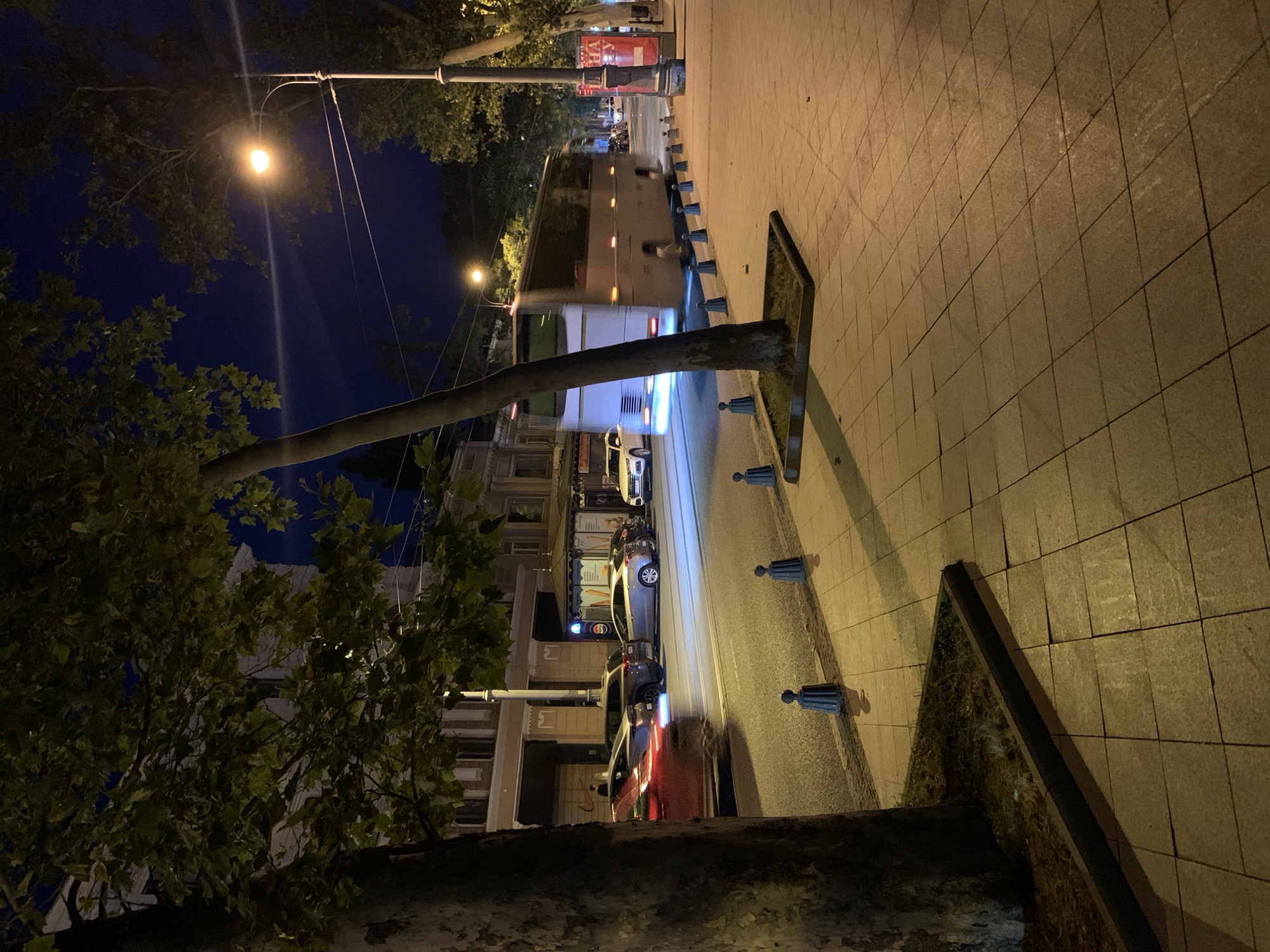The image depicts a street scene at night in Odesa, Ukraine. A large white bus is moving away from the camera on the right side of the road, with its headlights illuminated. Several cars are visible on the left side of the road, including a red car in motion and other parked vehicles. A sidewalk with patterned paving stones is in the foreground, bordered by a street and a tree. Streetlights illuminate the scene. Buildings with windows are visible in the background, and trees line the street.