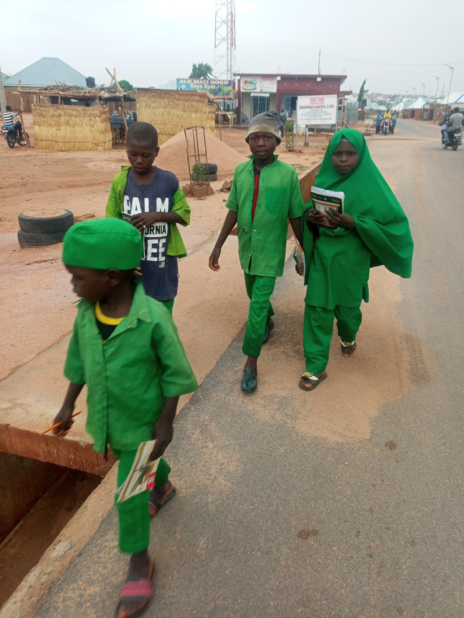 Four children are walking on a paved road in Tempure, Nigeria. The child closest to the camera is wearing a green cap, shirt, and pants, and holding a flat, rectangular object. To their right, a child wears a dark blue t-shirt and a green jacket. Another child further back wears a green shirt and pants. The last child, a girl, is wearing a green hijab and holds books. In the background, there are buildings, a sand pile, and parked tires. Motorbikes are visible on the road. A sign reads “EJSODAN HOTEL LTD.”