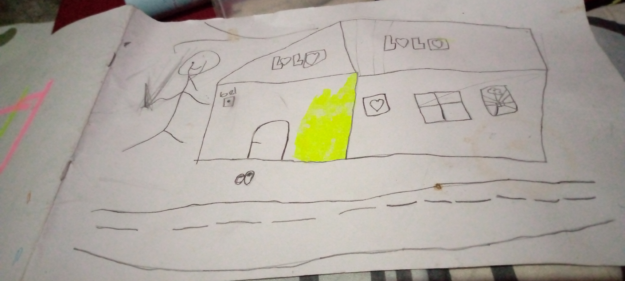 A child's drawing on white paper depicts a simplistic house with a door and two windows. The house has "LOLO" written on its roof in block letters with hearts interspersed. A stick figure stands to the left of the house. Below the house, a road is drawn with dashed lines. The drawing is not in the city of Sumberwono, Indonesia.