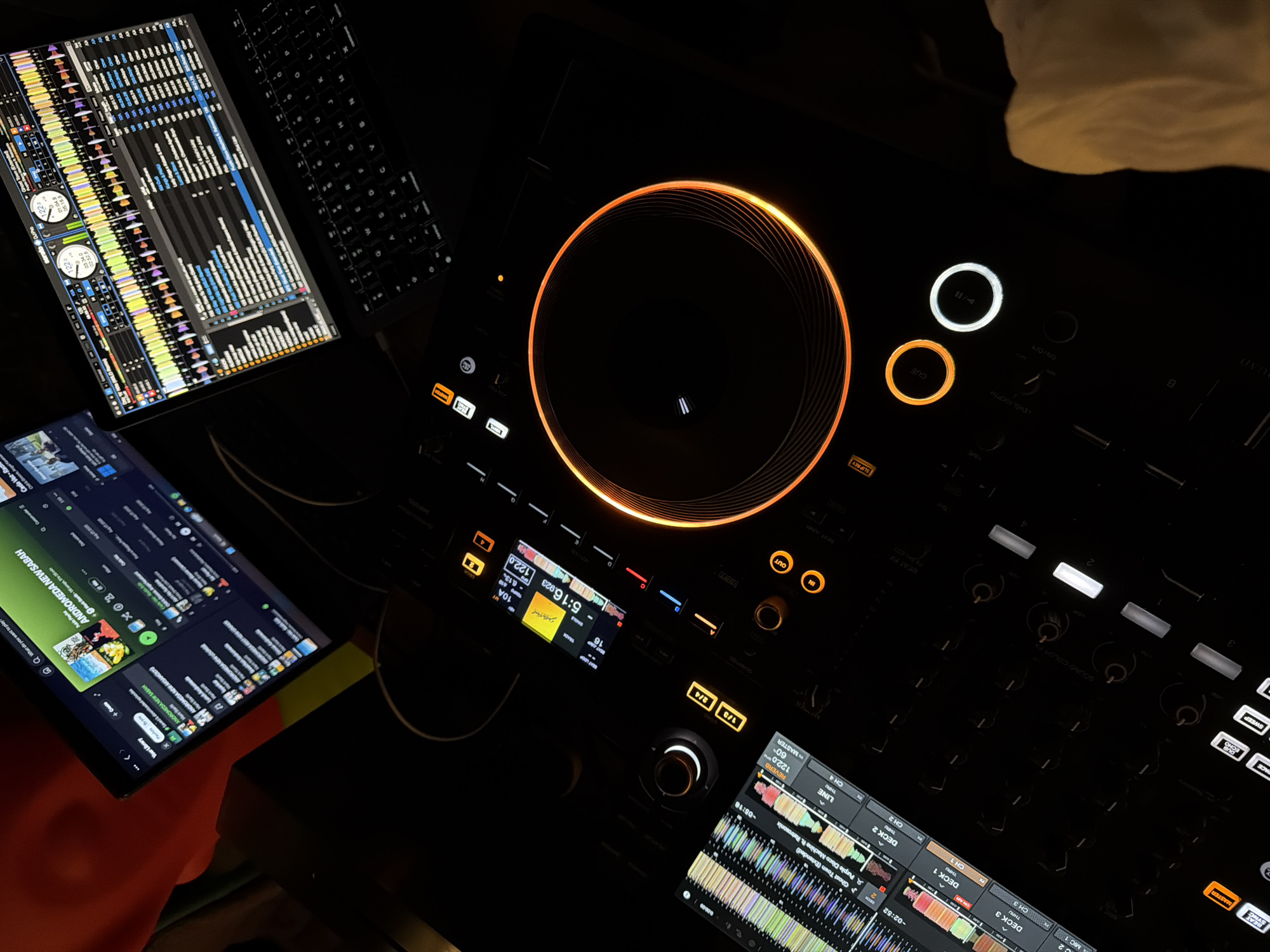 The image depicts a DJ setup in a dimly lit environment. The central object is a DJ controller with an illuminated jog wheel and various buttons and knobs. Flanking the controller are two screens displaying audio waveforms and other DJ software interfaces. One screen shows a music library, including the song "ANDROMEDA NEW SABAN". A keyboard is partially visible near one of the screens. The close-up nature of the image and the specific details of the DJ equipment suggest a focus on the technical aspects of DJing. There is no information in the image to infer the location of Budva, Montenegro.