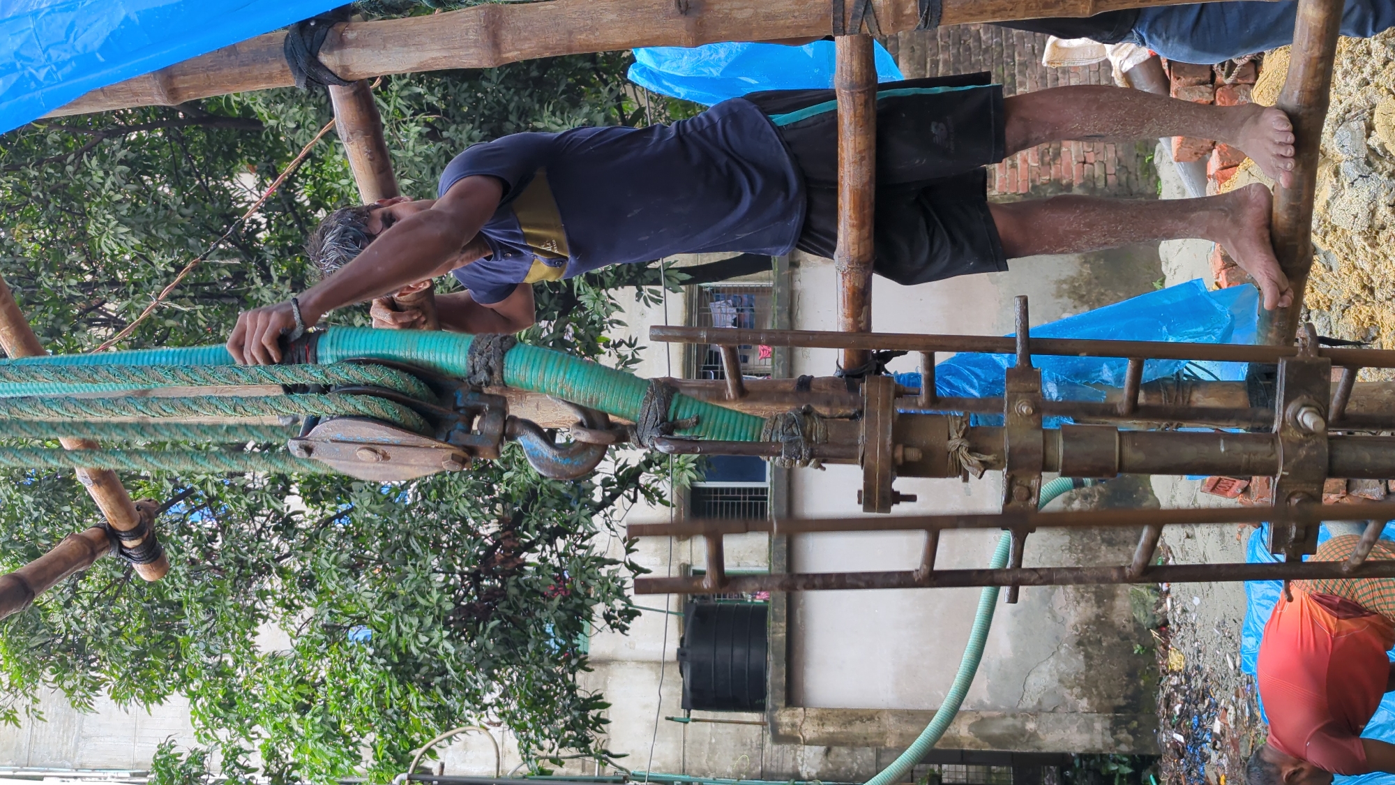 A man is working with a drilling rig in Dhaka, Bangladesh. The man, wearing a dark blue and yellow t-shirt and black shorts, is barefoot and standing on a wooden beam, gripping a green hose that is part of the drilling equipment. The hose is attached to a pulley system suspended from bamboo scaffolding. In the background, a building with windows is visible, along with trees and a black water tank. Another man, wearing a red shirt and checkered pants, is visible in the lower left corner, also working near the drilling rig. Blue tarps are draped over some of the equipment, and debris is scattered on the ground. The overall scene suggests construction or repair work in an outdoor urban environment.
